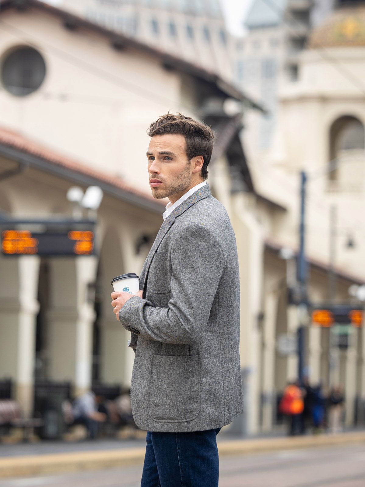 Stylish young man in gray blazer holds coffee cup while standing at an urban train station platform.