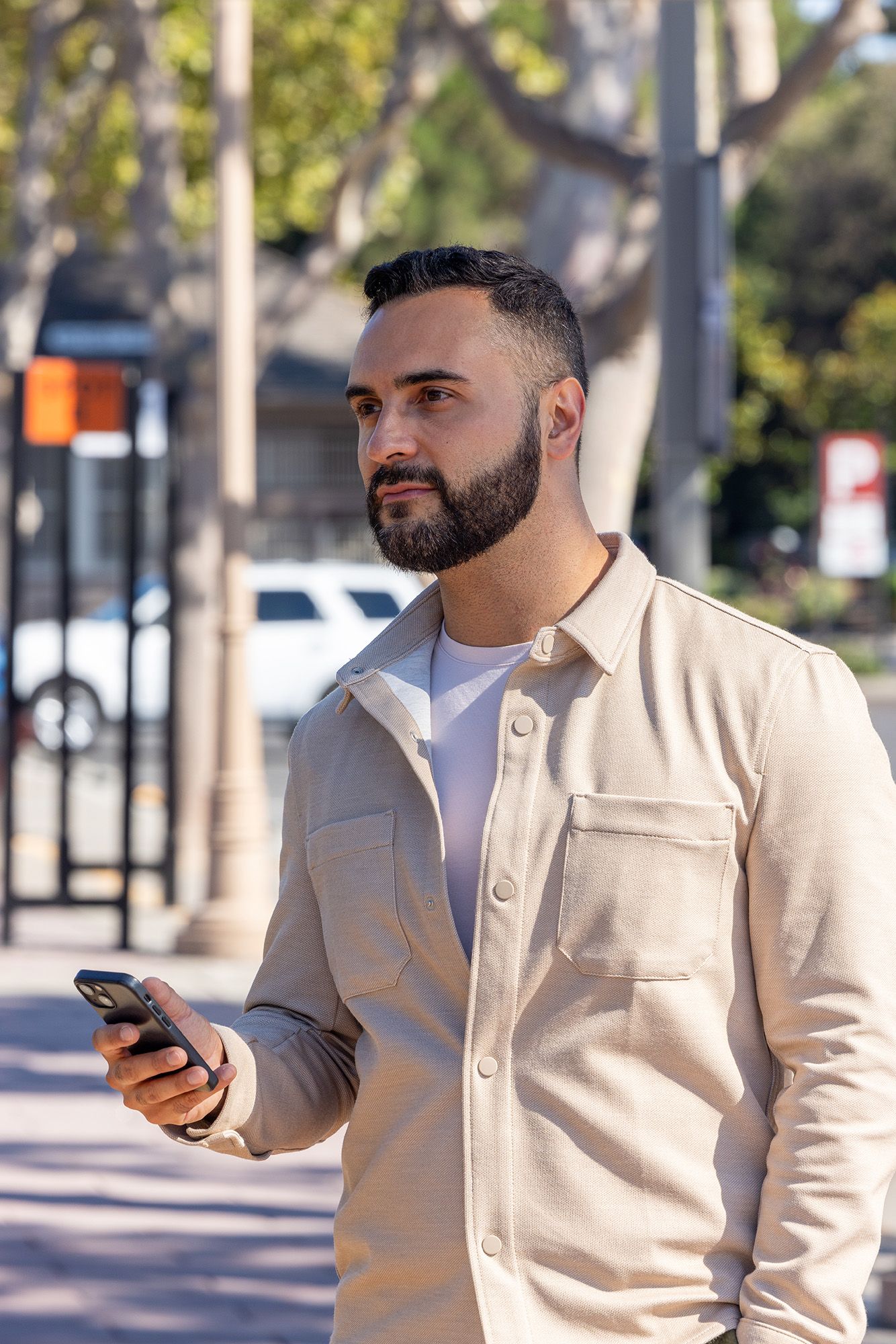 Bearded man in beige jacket holding smartphone while standing on sunny urban sidewalk lined with trees.