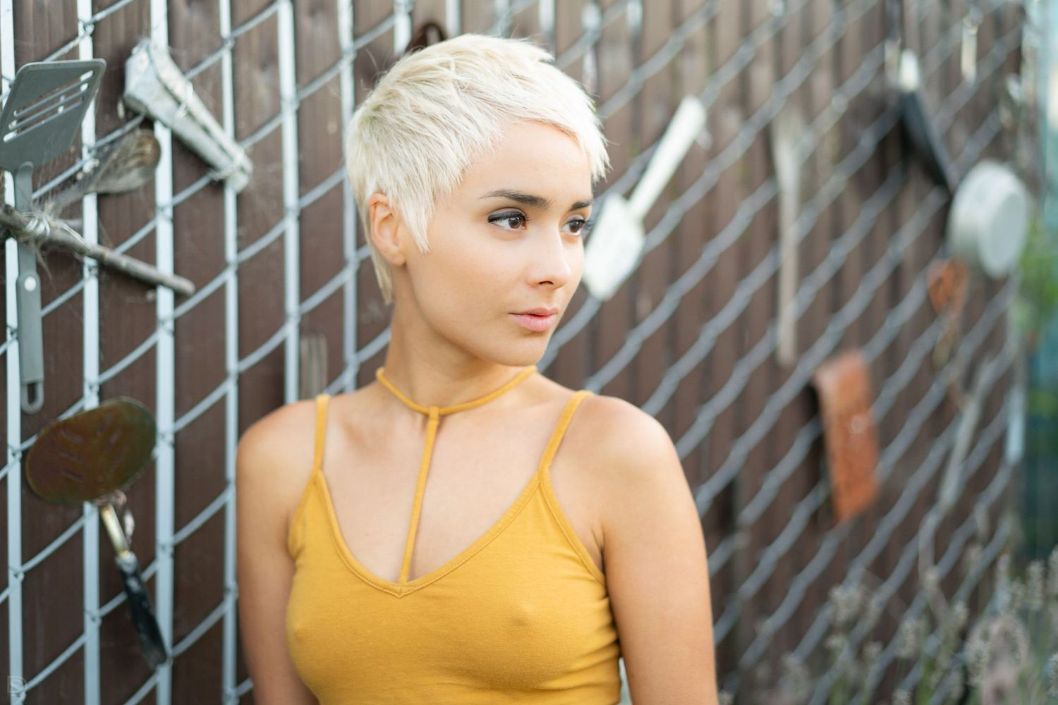 Young woman with platinum pixie cut in yellow tank top leaning against a chain-link fence outdoors.