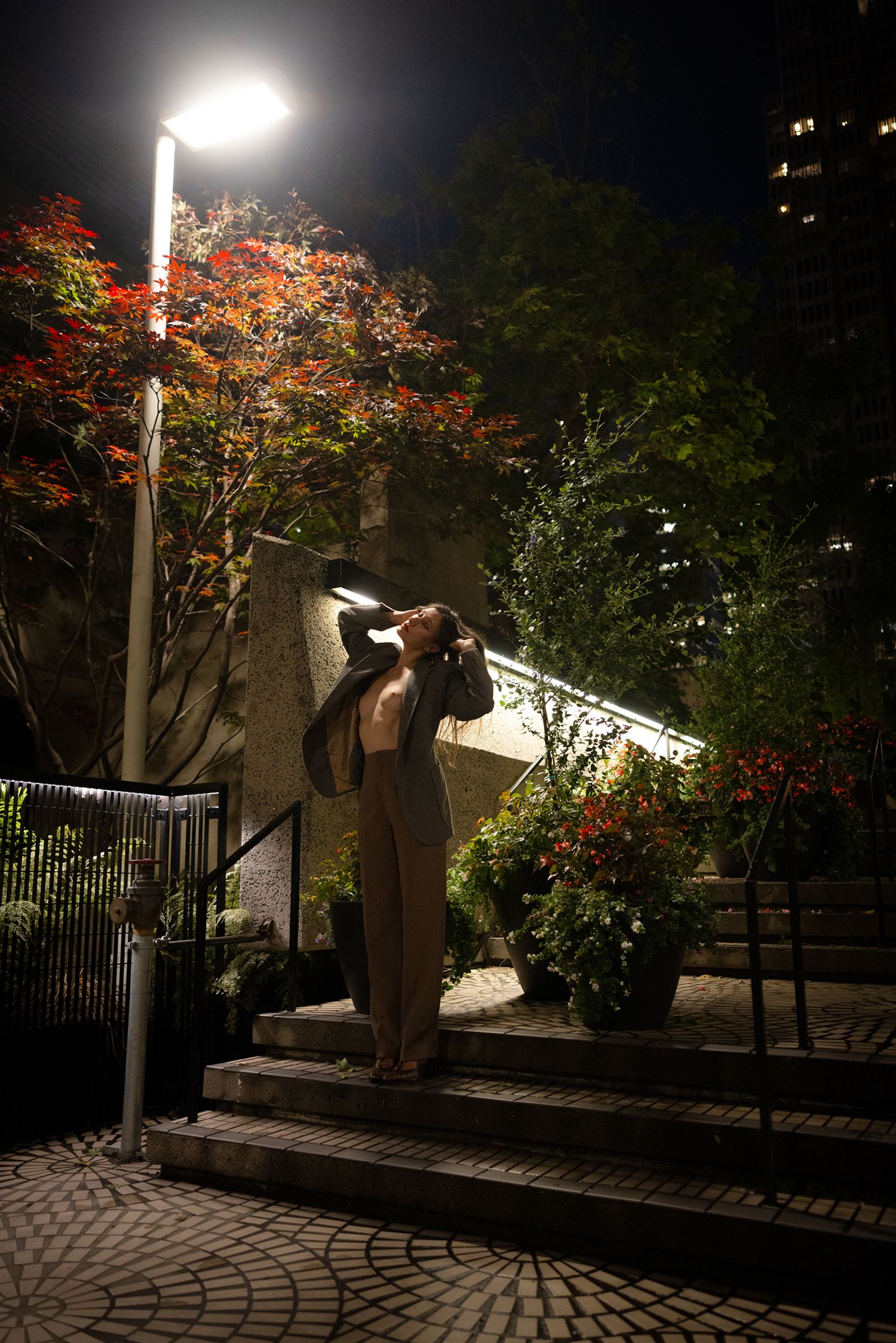 Woman standing under a street lamp at night beside colorful autumn trees and flower-lined steps.