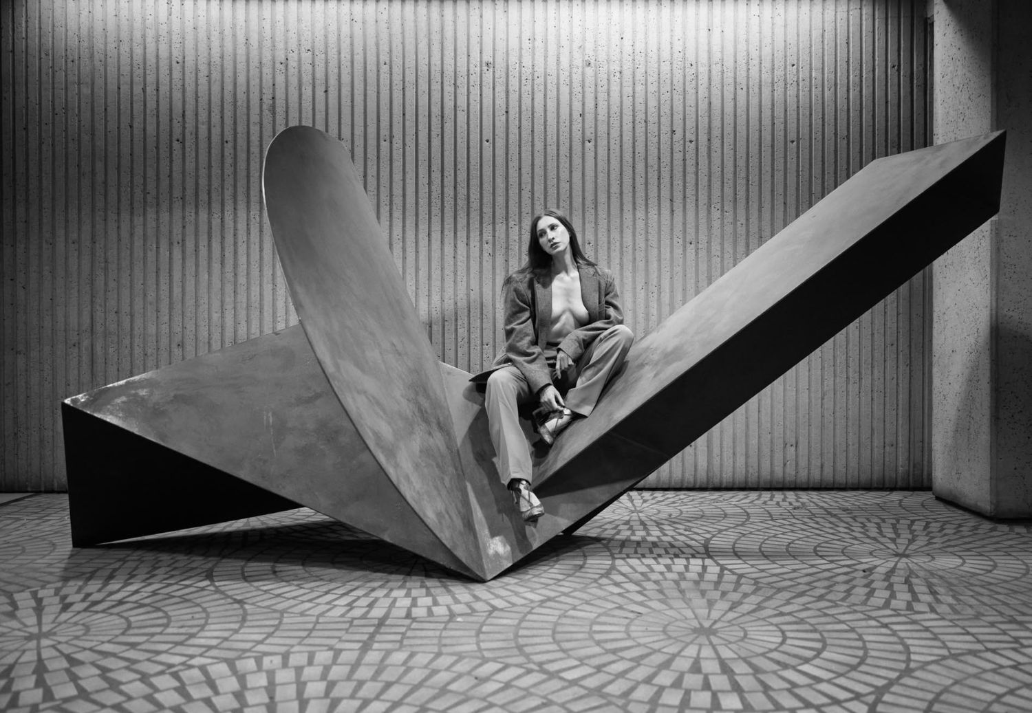 Woman in blazer sitting on large abstract metal sculpture in black and white photo.
