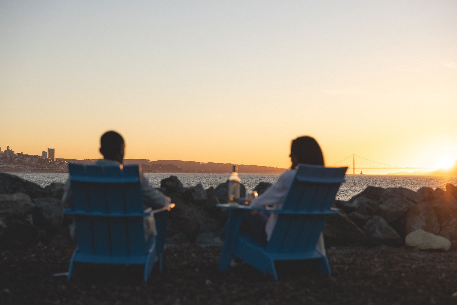 Two people sitting in blue Adirondack chairs watching a golden sunset over the water on a rocky beach.