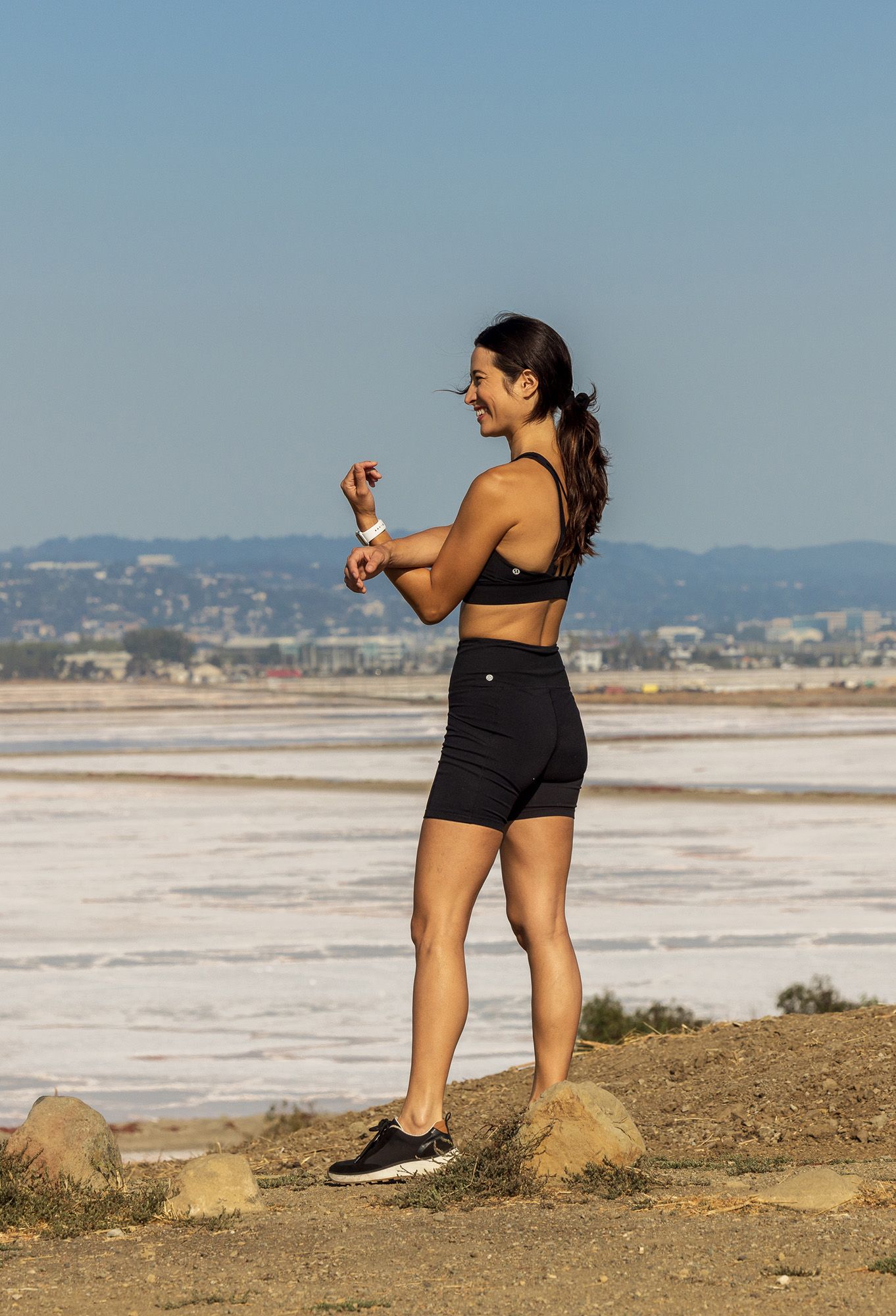 Woman in black athletic wear checks smartwatch outdoors near coastal salt flats on a sunny day.