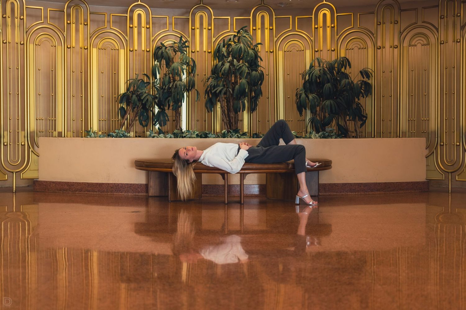 Woman lying on a wooden bench in an ornate golden Art Deco lobby with indoor plants.