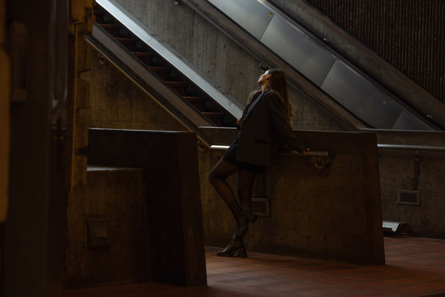 Lone figure leaning against concrete wall near escalator in dimly lit urban underground setting at night.
