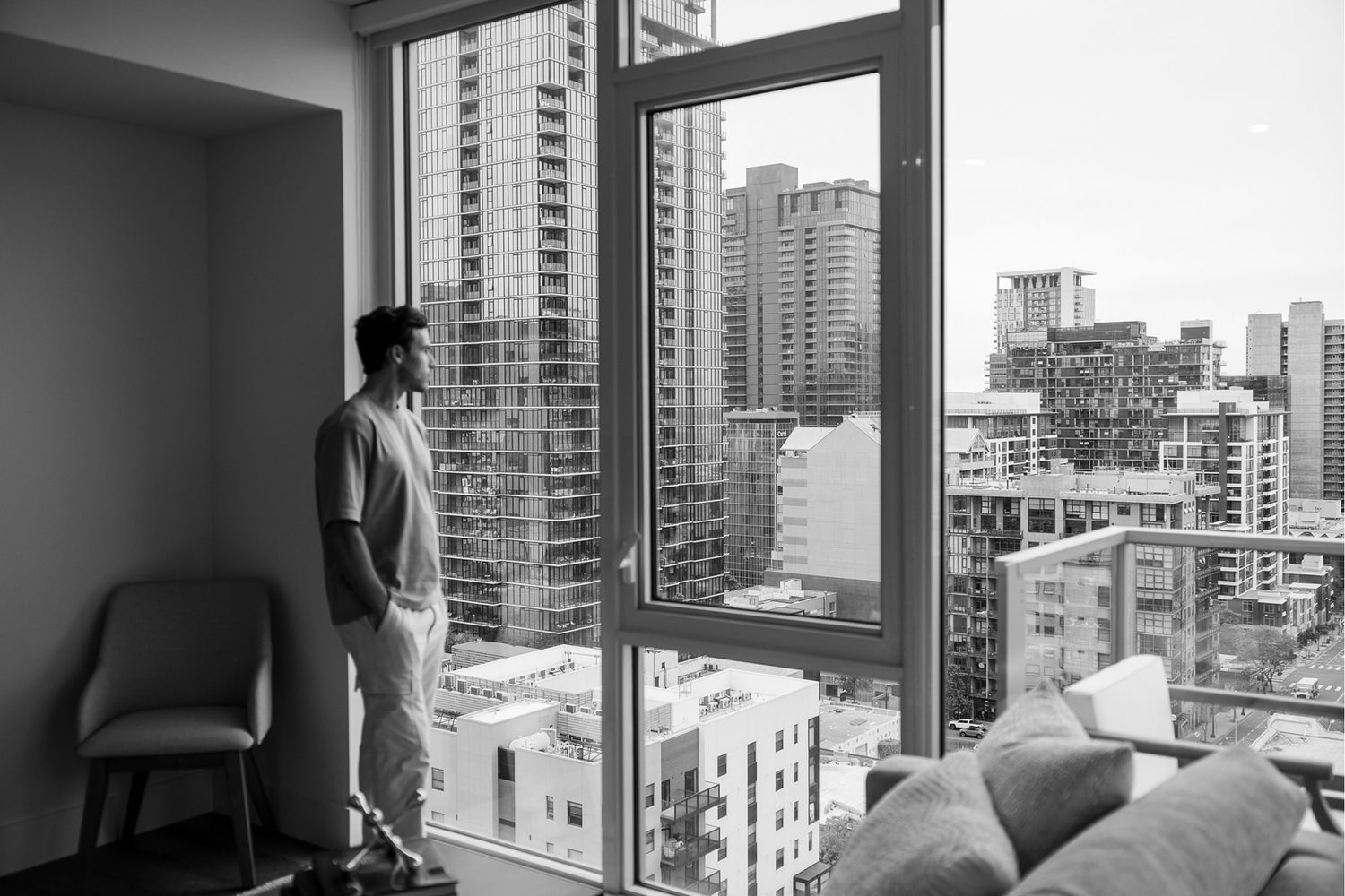 Young man in casual outfit stands by floor-to-ceiling windows overlooking a dense urban cityscape in black and white.