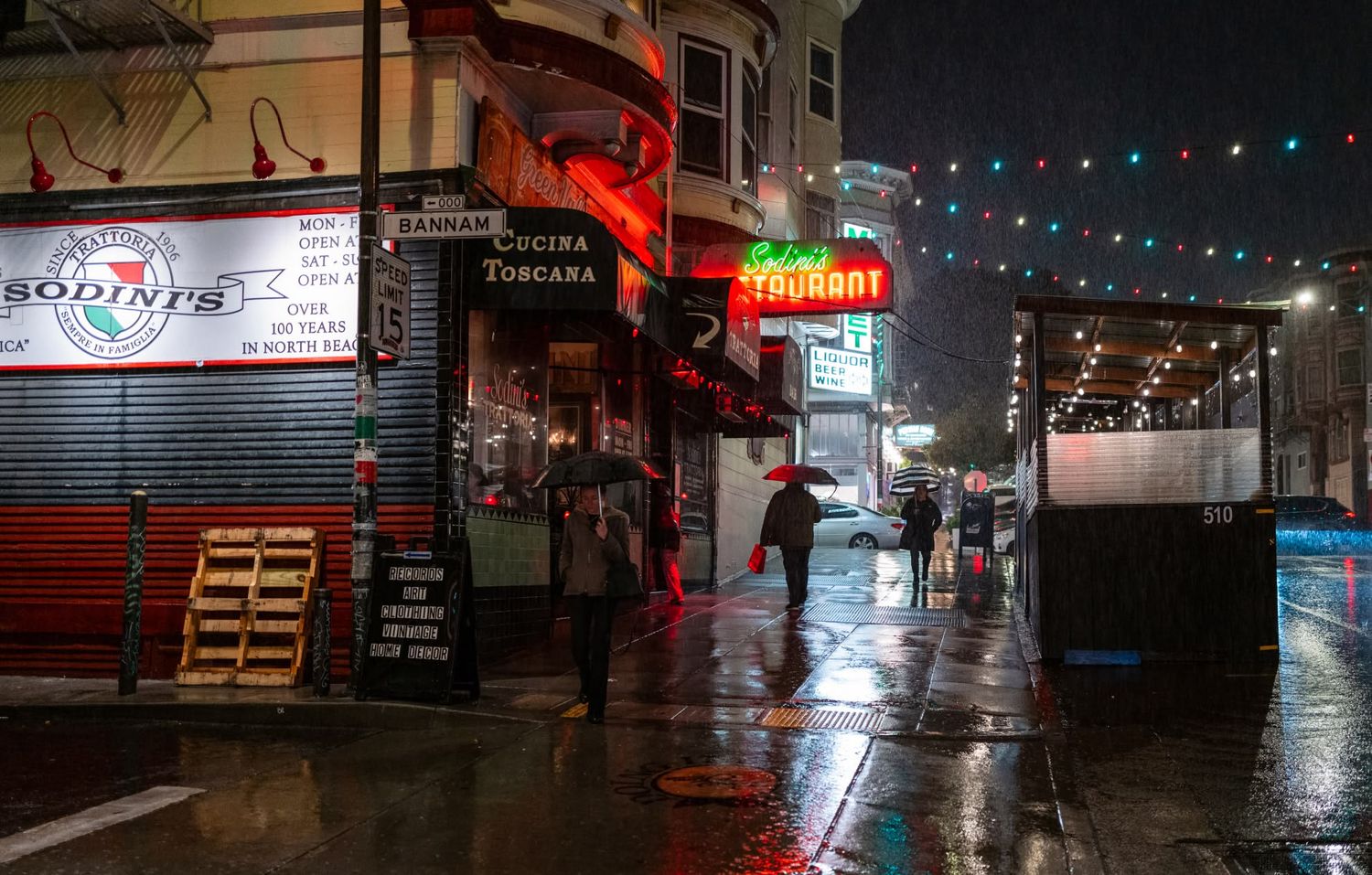 Rainy night street scene in San Francisco with neon restaurant signs, umbrellas, and wet reflective sidewalks.