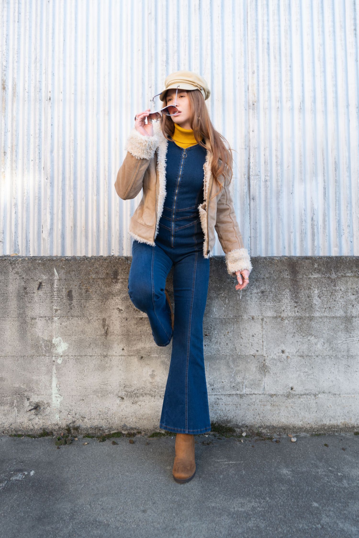 Stylish woman in flare jeans, shearling jacket, and newsboy cap posing against a corrugated metal wall.