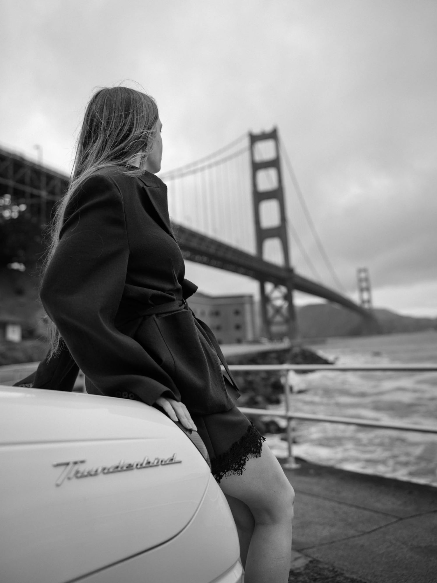 Black and white photo of a woman leaning on a classic car near the Golden Gate Bridge in San Francisco.