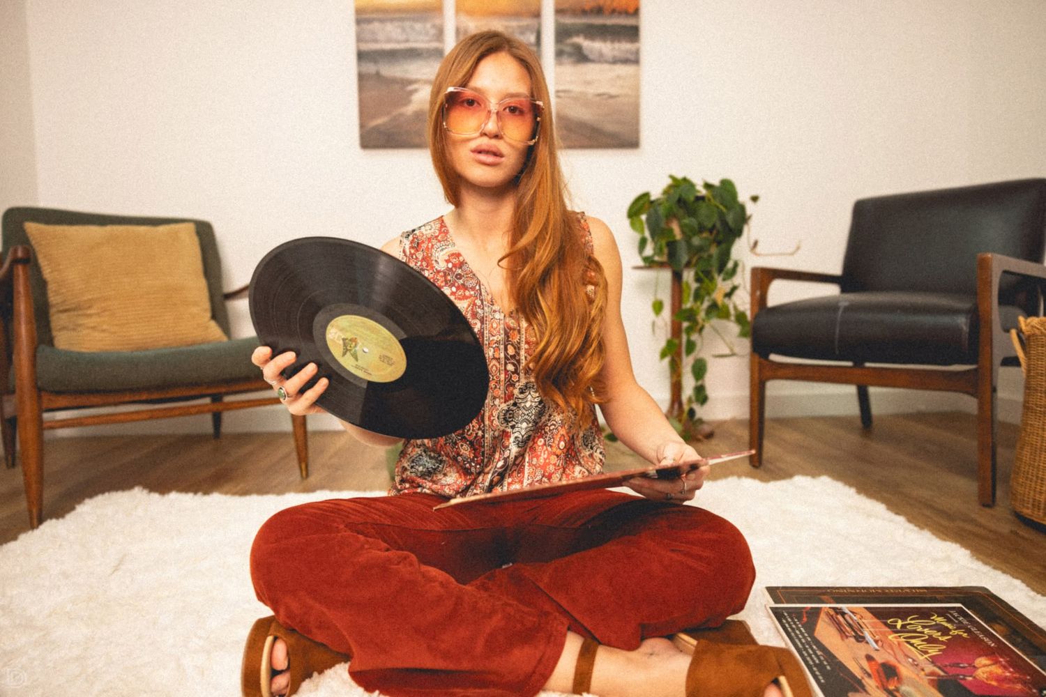 Retro-styled woman sitting cross-legged on white rug holding a vinyl record wearing red velvet flares and pink sunglasses.