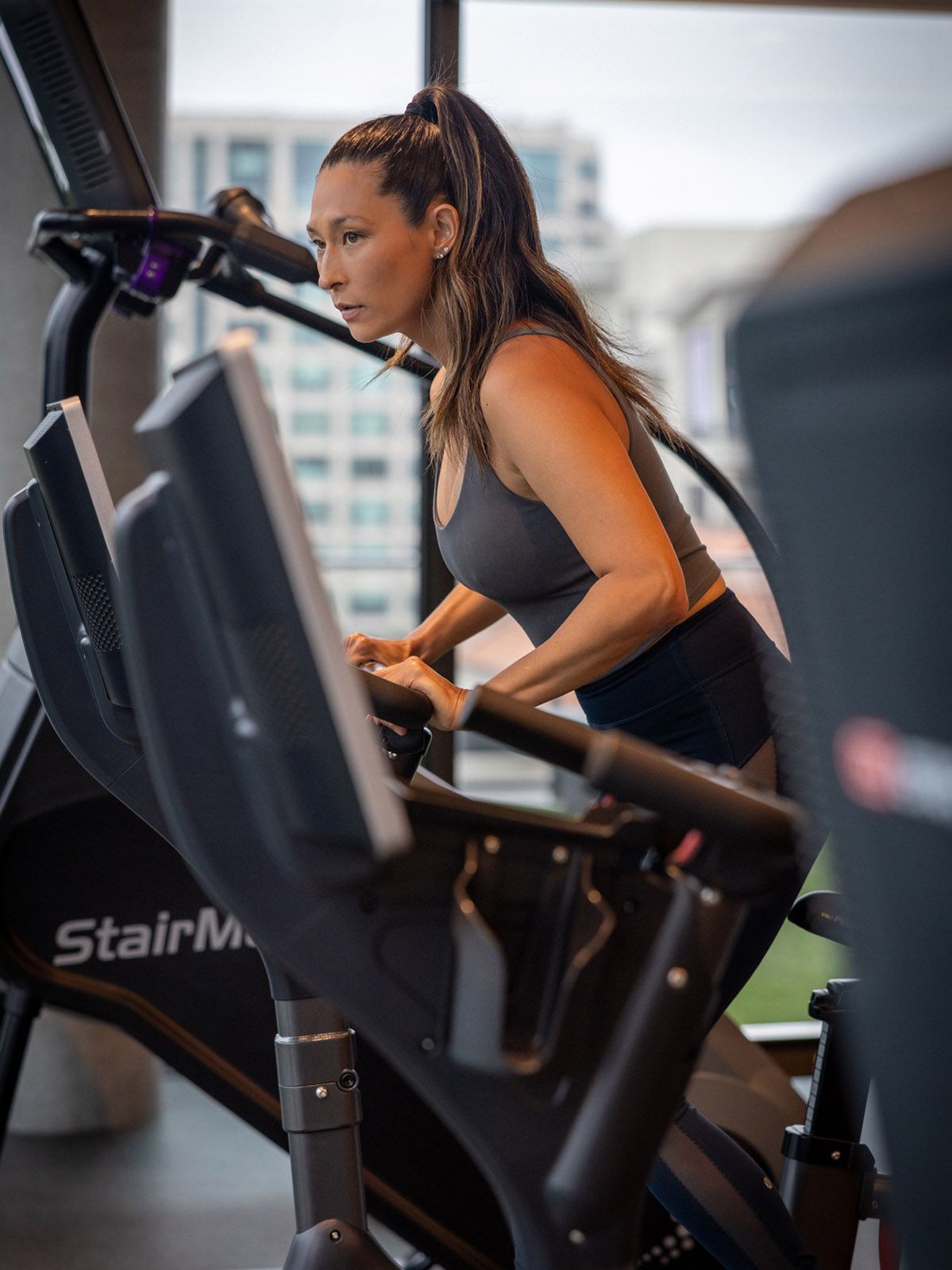 Focused woman with ponytail exercises intensely on a StairMaster machine in a modern gym with city views.