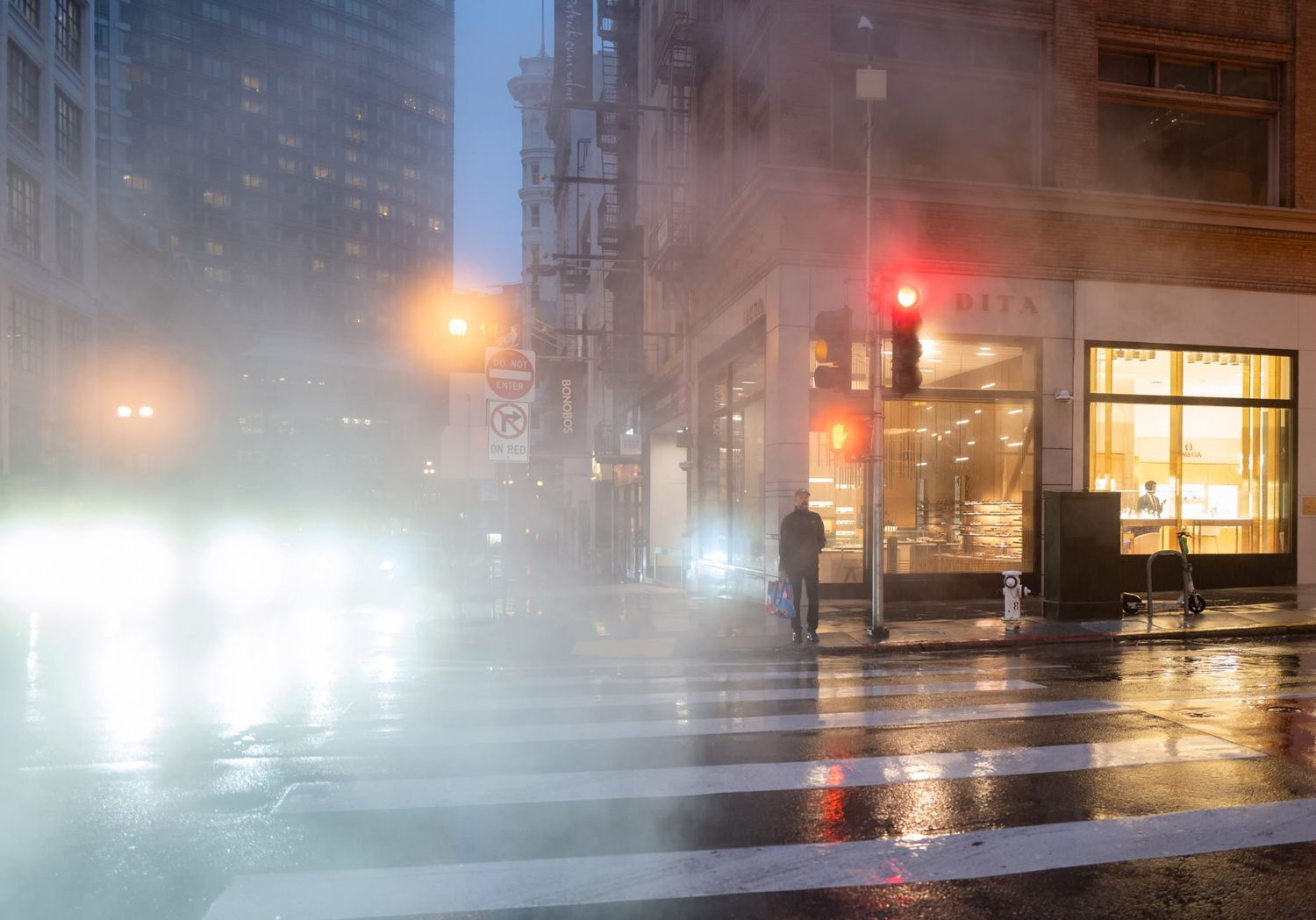 Foggy rainy urban intersection at night with steam, glowing streetlights, red traffic signal, and a lone pedestrian.