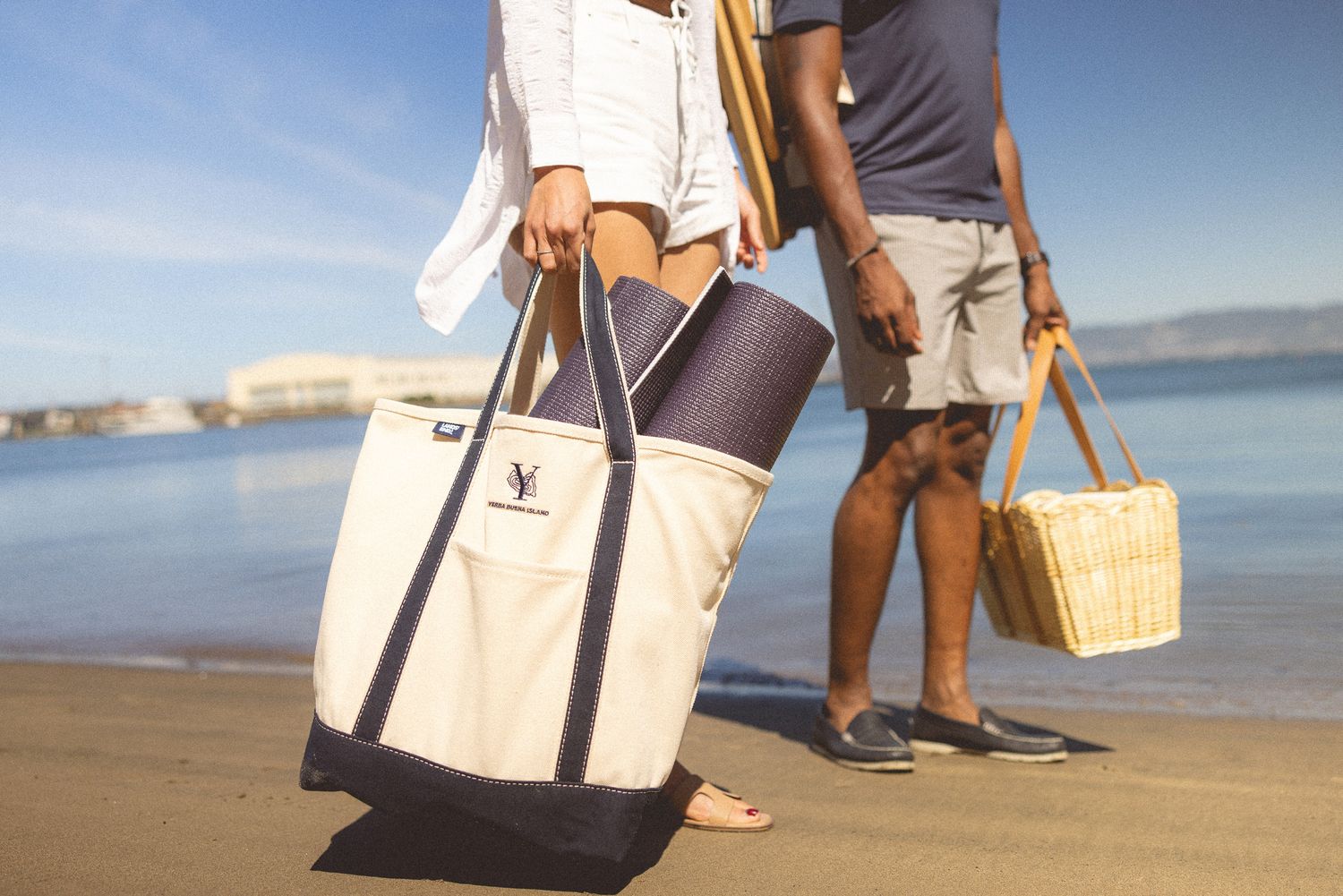 Couple on beach carrying navy and white tote bag and wicker picnic basket on sunny day.