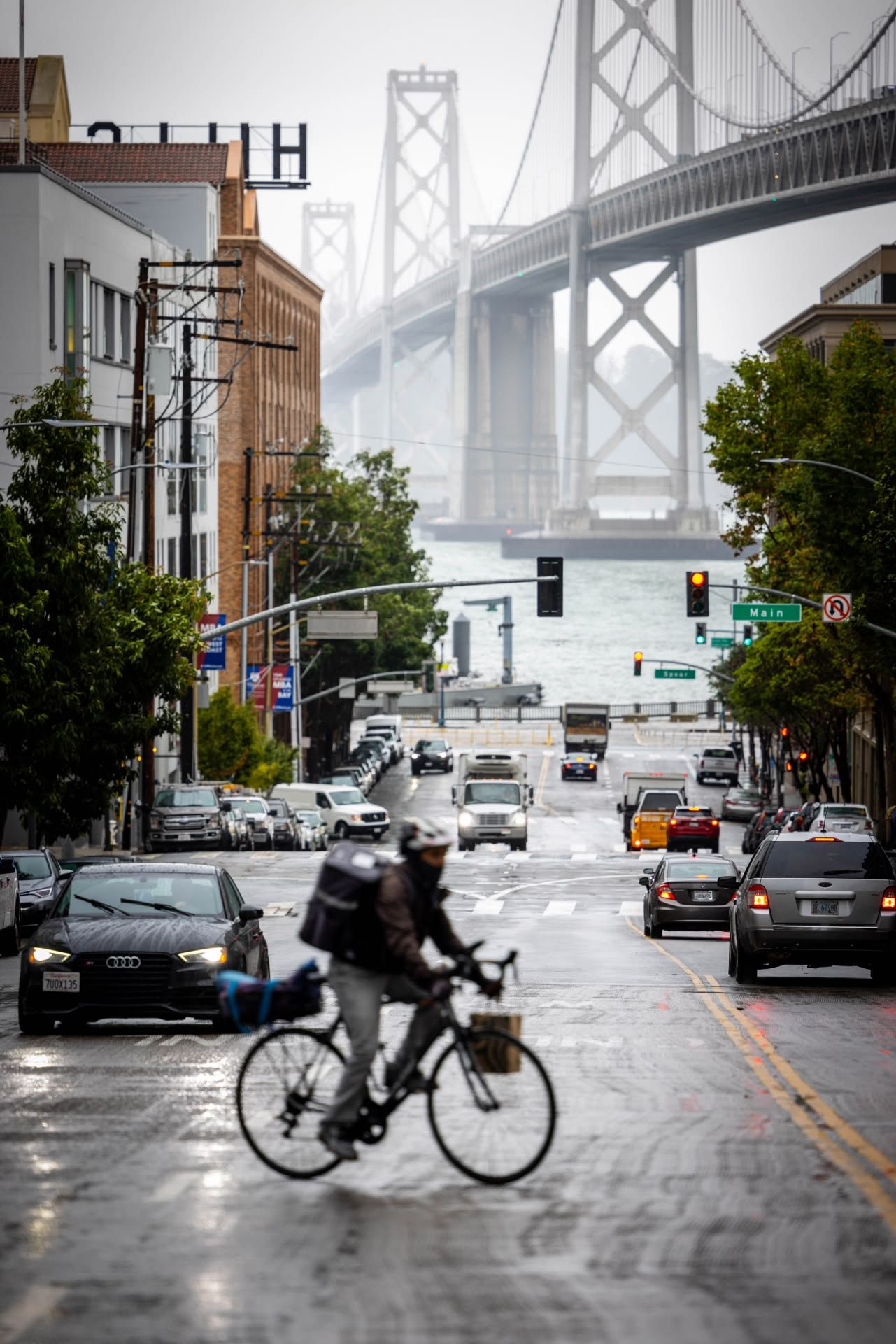 Cyclist crossing a wet street in San Francisco with the Bay Bridge visible in the foggy background.