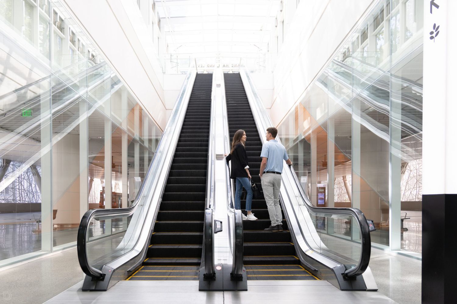 Two people riding escalators in a bright modern building with large windows and high ceilings.