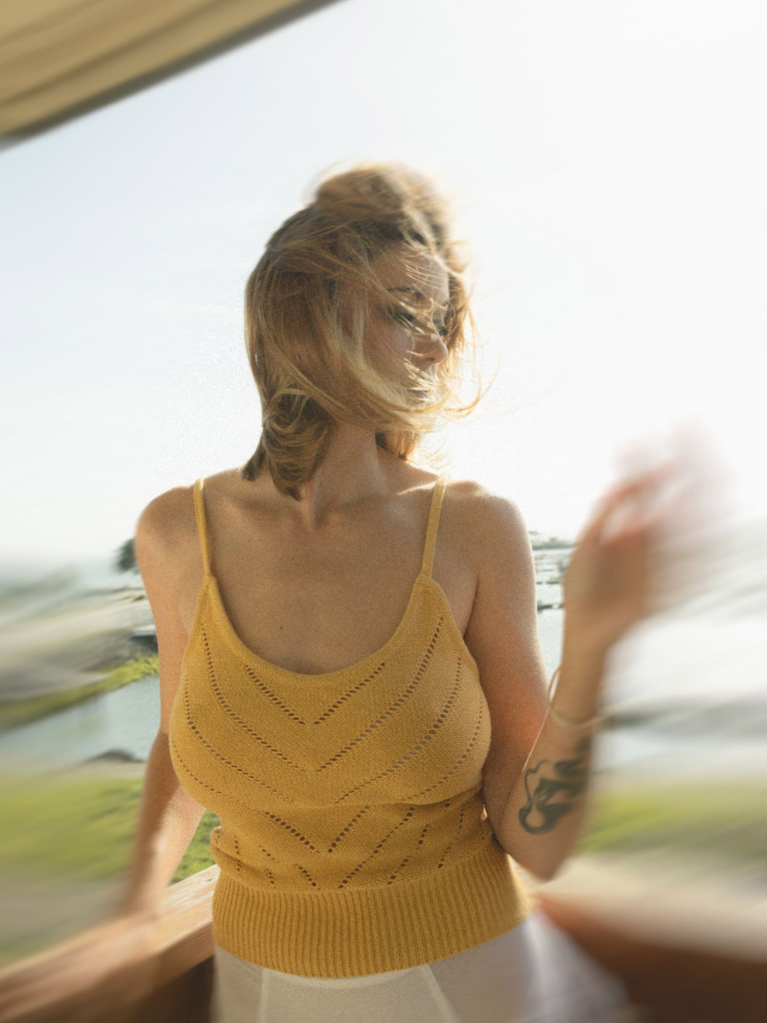 Woman in yellow knit crochet tank top standing on sunny balcony with wind-blown hair.