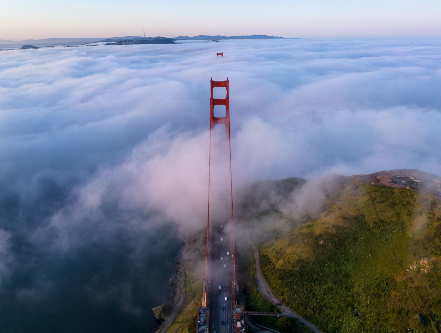 Aerial view of Golden Gate Bridge tower rising above thick white fog at dawn.