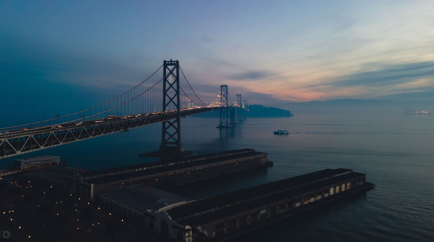 Dramatic dusk aerial view of San Francisco Bay Bridge with glowing lights reflecting on calm bay waters.