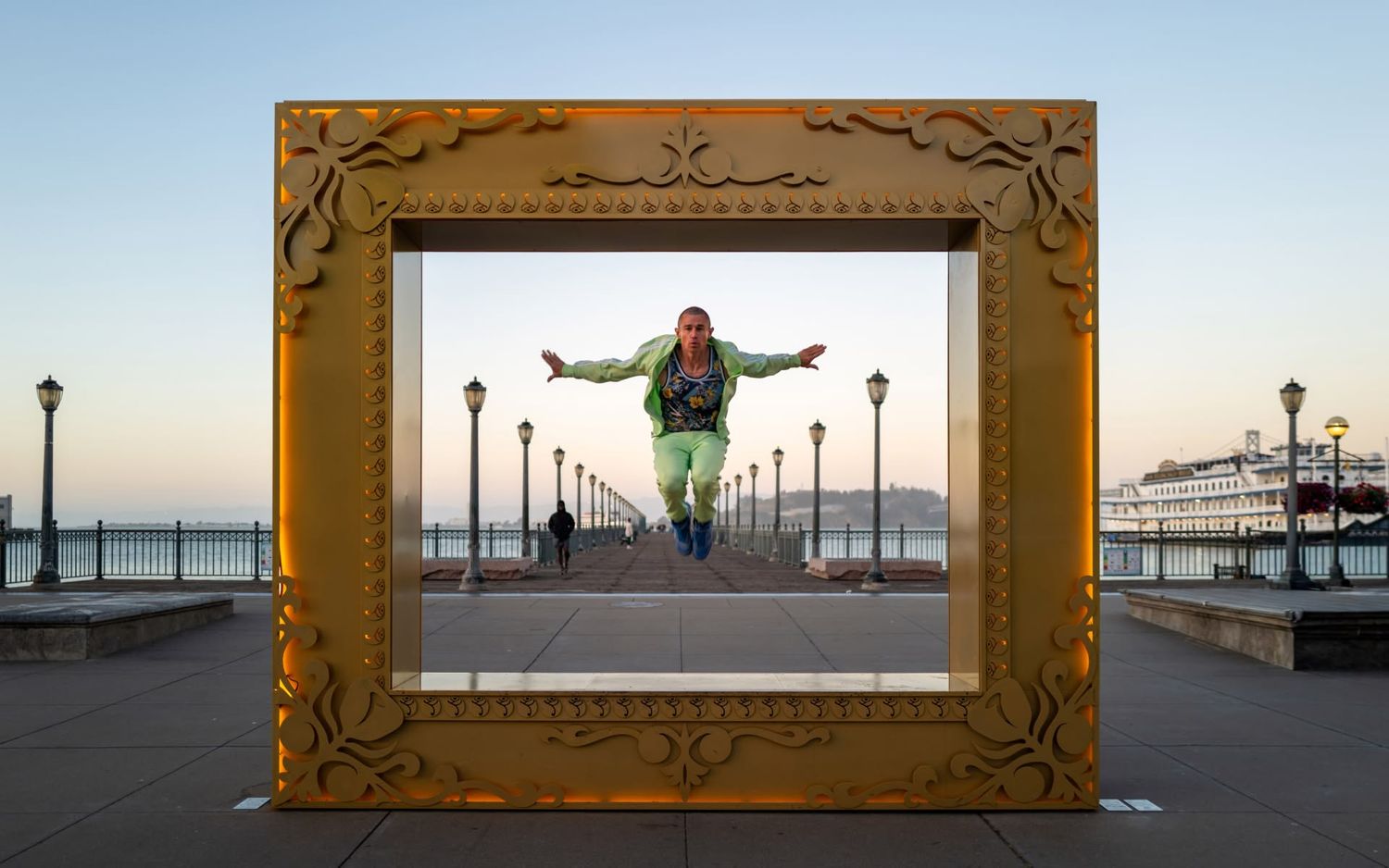 Person jumping inside a large ornate gold picture frame on a waterfront promenade at sunset.