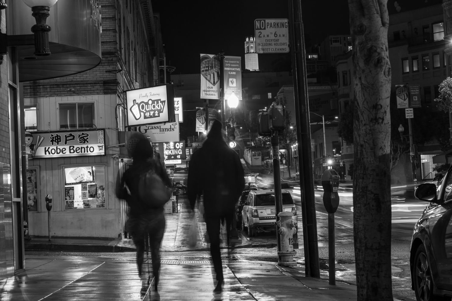 Black and white nighttime street scene with two people walking past Kobe Bento restaurant on a wet urban sidewalk.