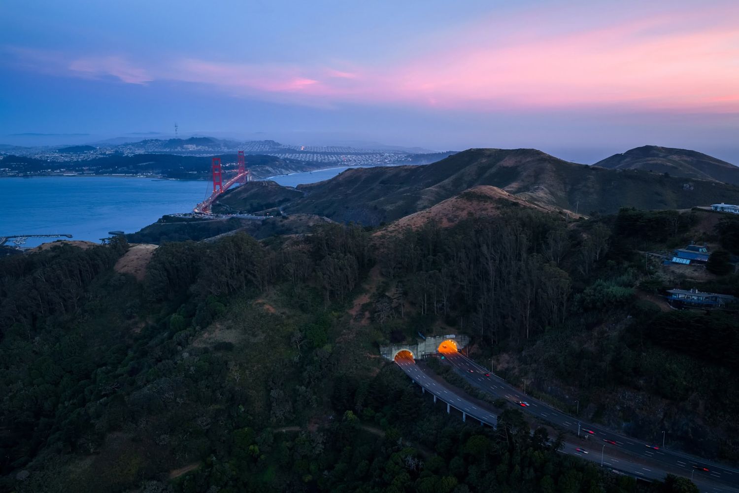 Aerial dusk view of Golden Gate Bridge and rolling hills with highway tunnels glowing under a pink and purple sky.