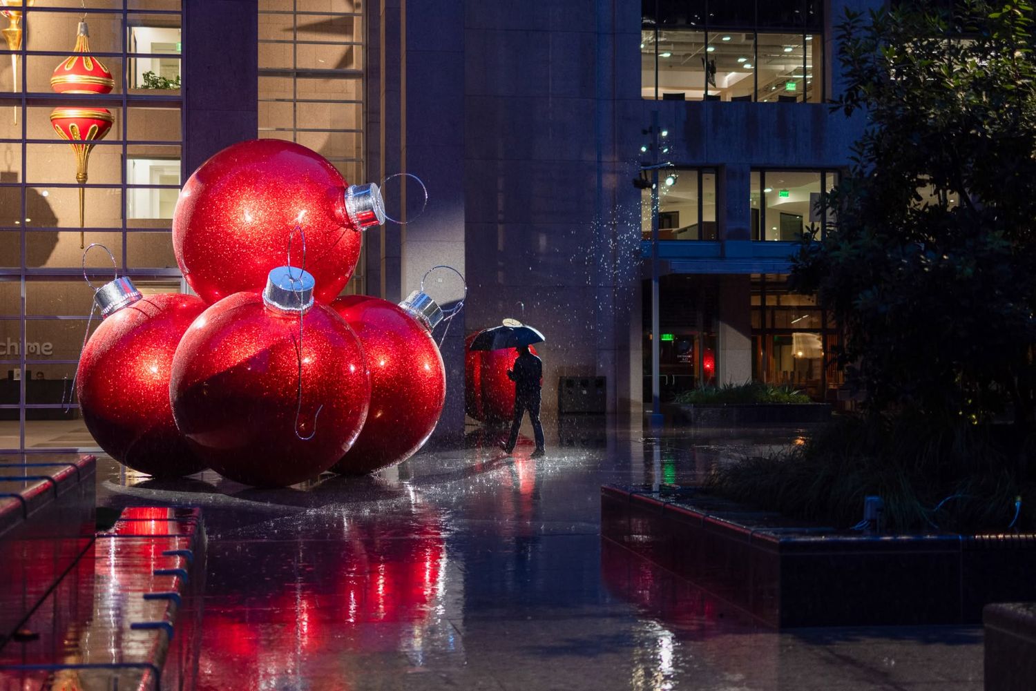 Giant red Christmas ornaments displayed outside a modern building at night, reflected on wet pavement in the rain.