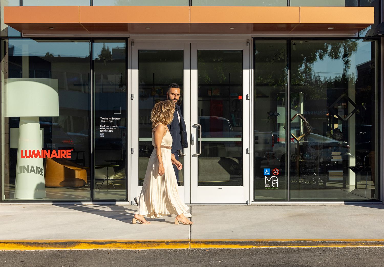 A couple walks hand in hand outside the Luminaire store entrance on a sunny day.