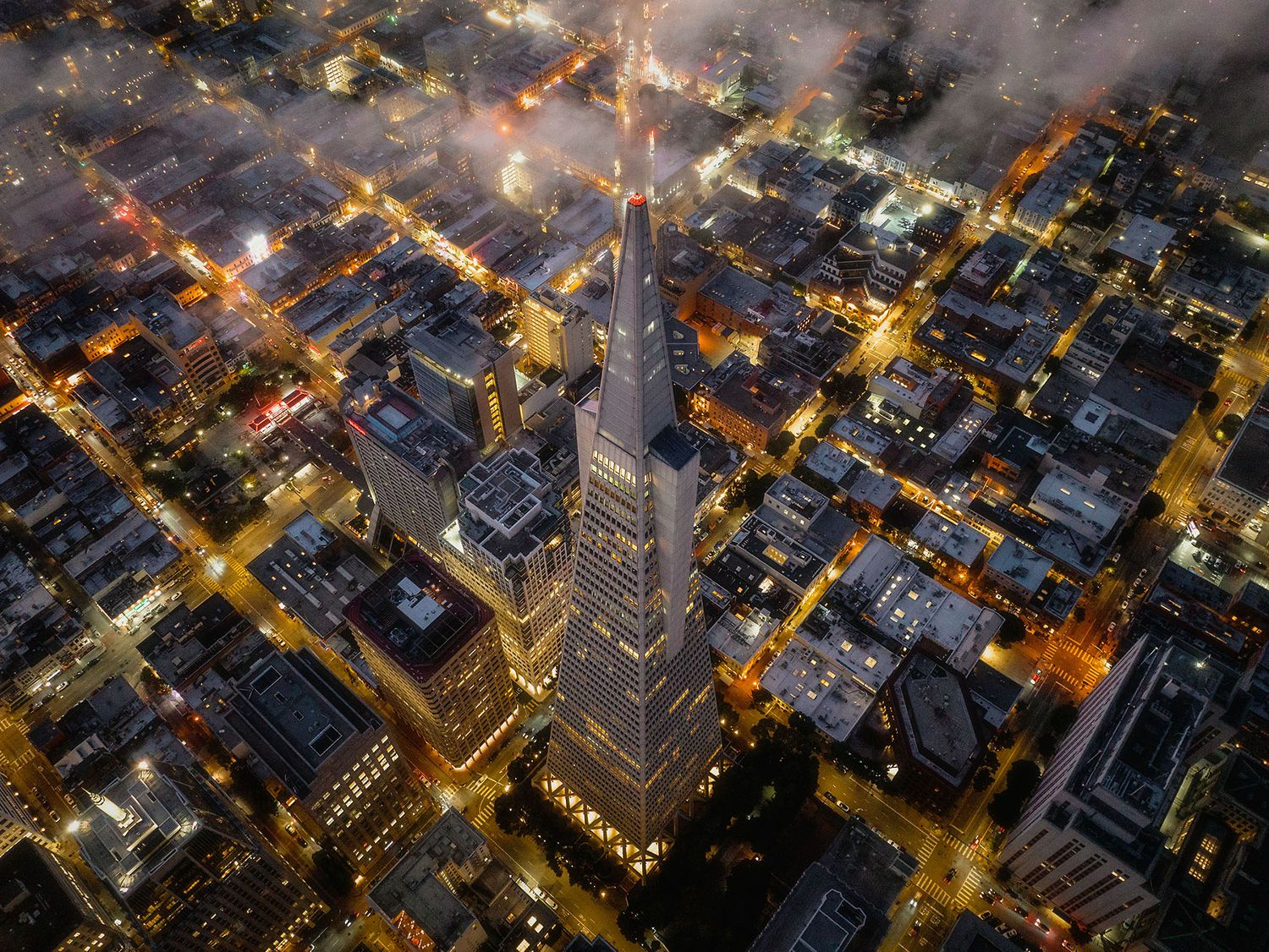 Aerial night view of Transamerica Pyramid surrounded by glowing city streets and misty fog in San Francisco.