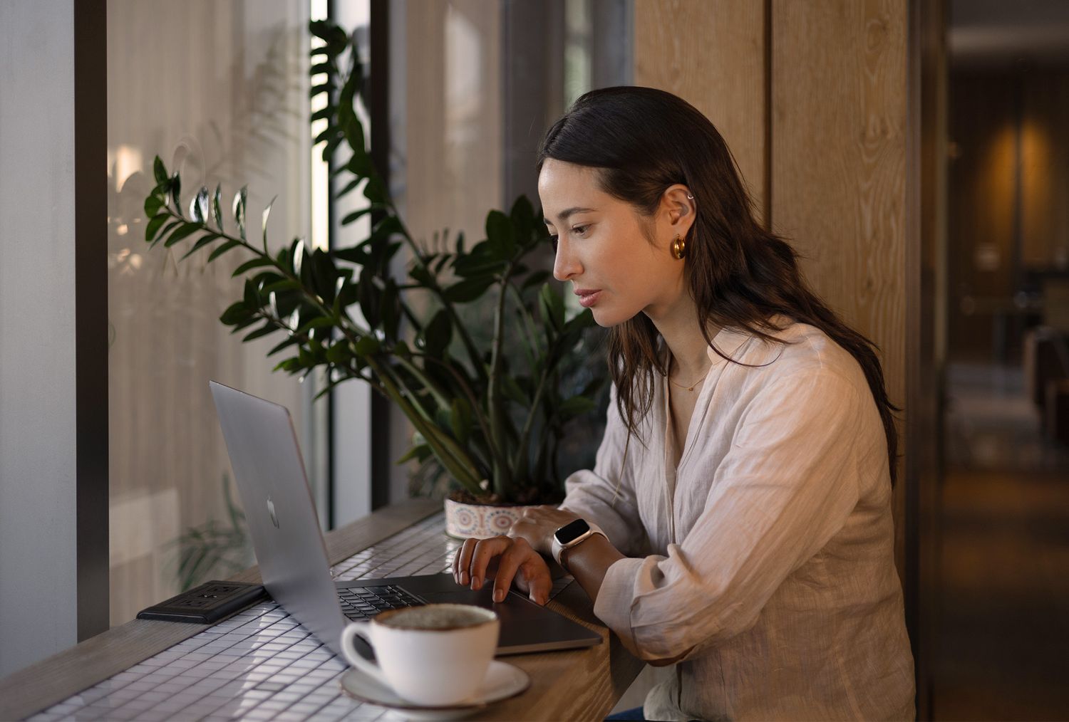 Young woman working on laptop at cafe table with coffee cup and green plants in background.
