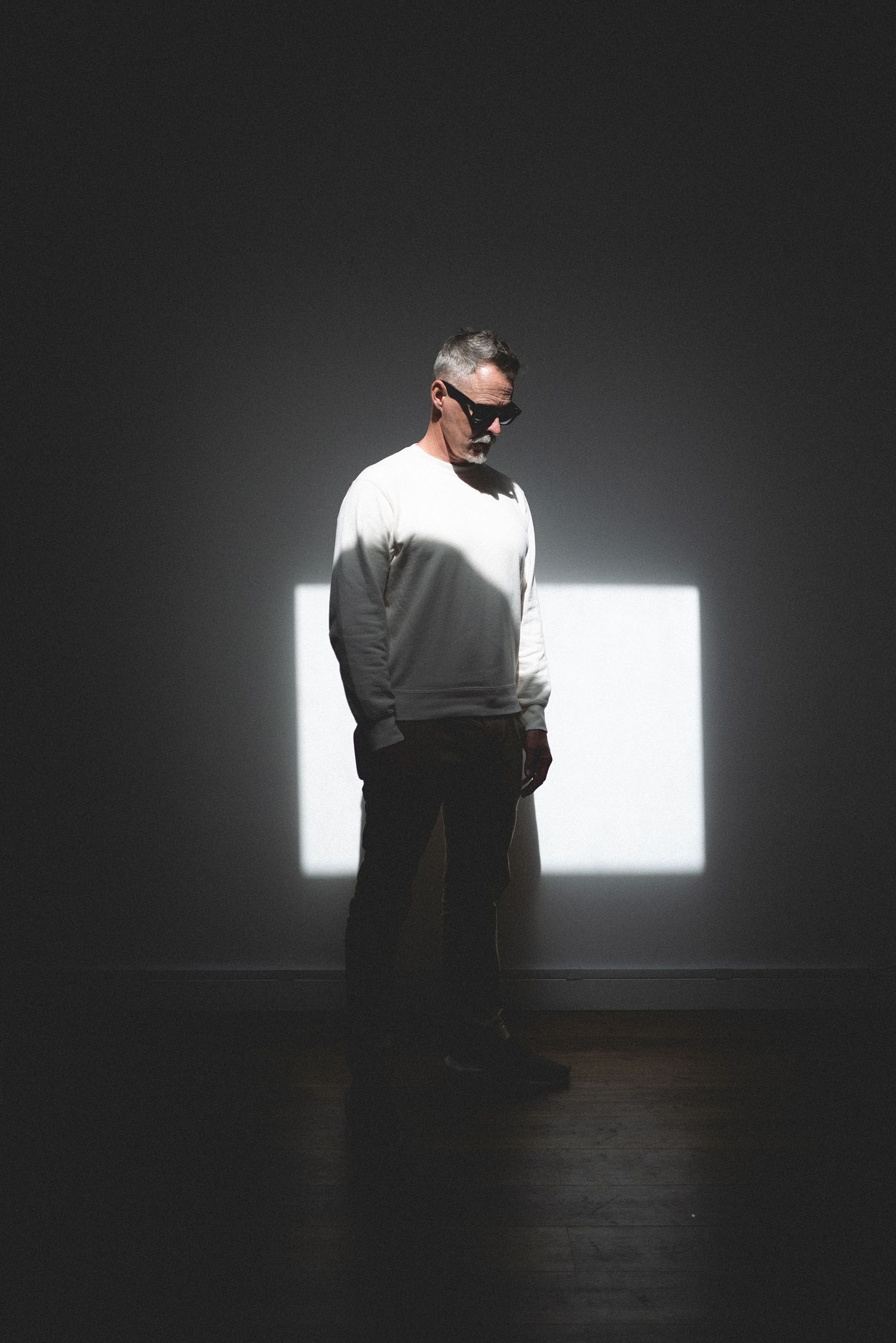 Middle-aged man in white sweatshirt standing in dark room with square light projection behind him.
