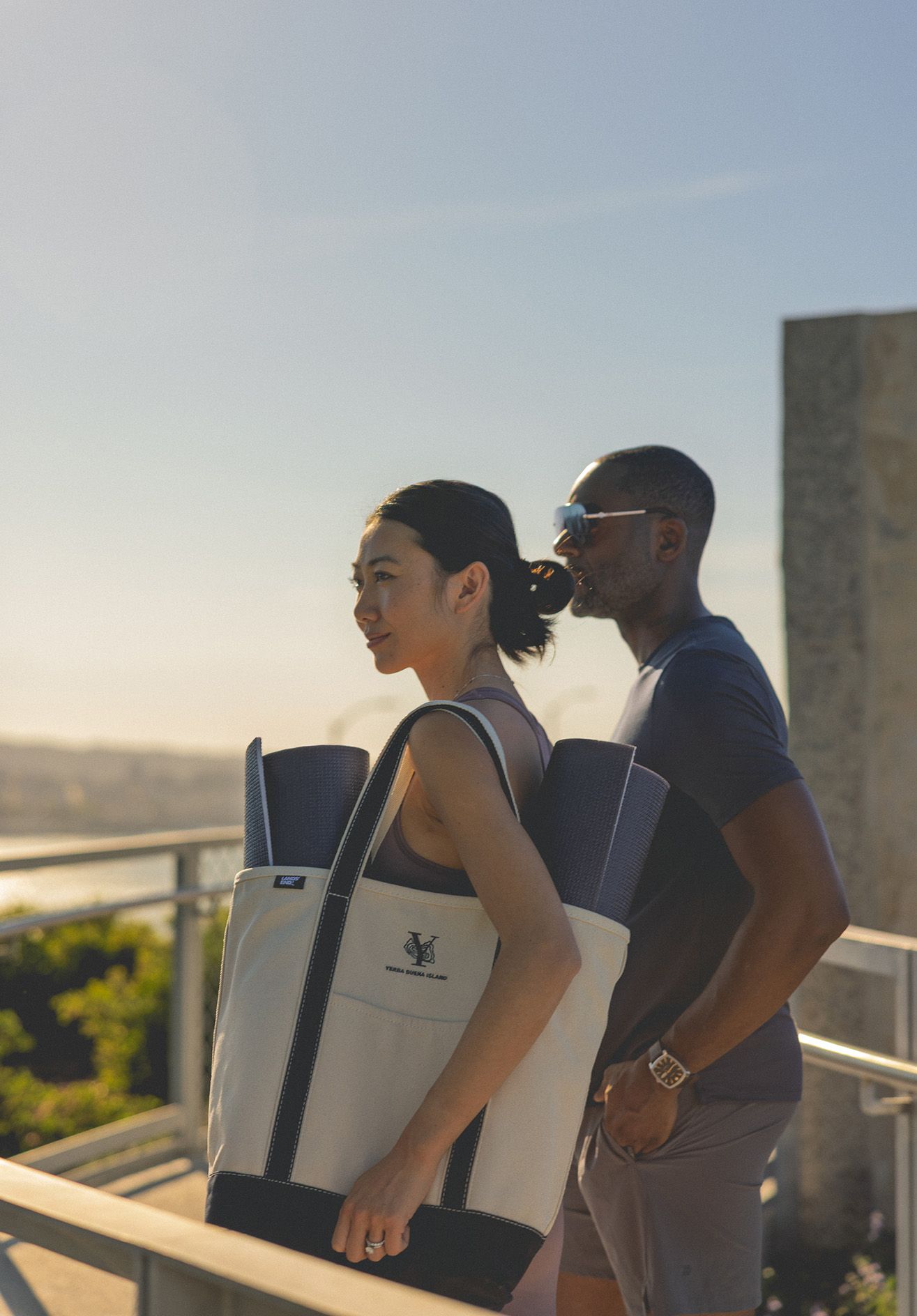 Two people carrying matching canvas tote bags with yoga mats outdoors at golden hour overlooking a scenic waterfront.
