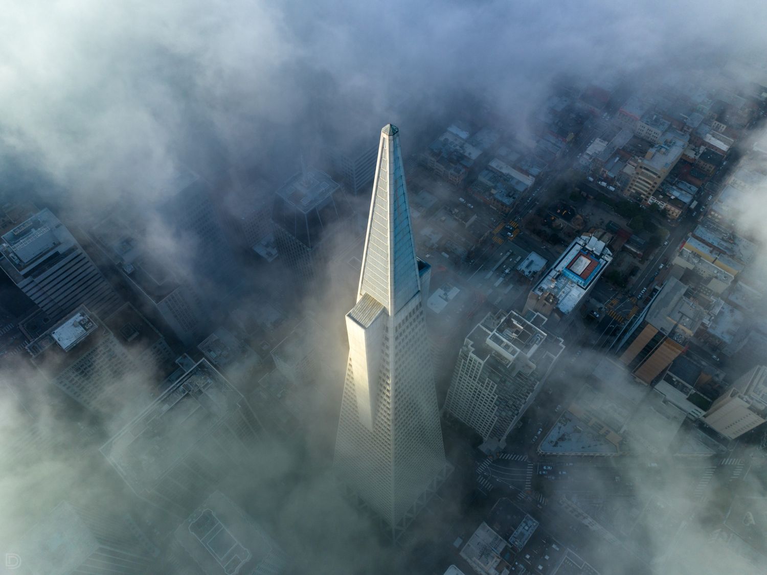 Aerial view of San Francisco Transamerica Pyramid skyscraper piercing through dramatic fog and clouds over the city.