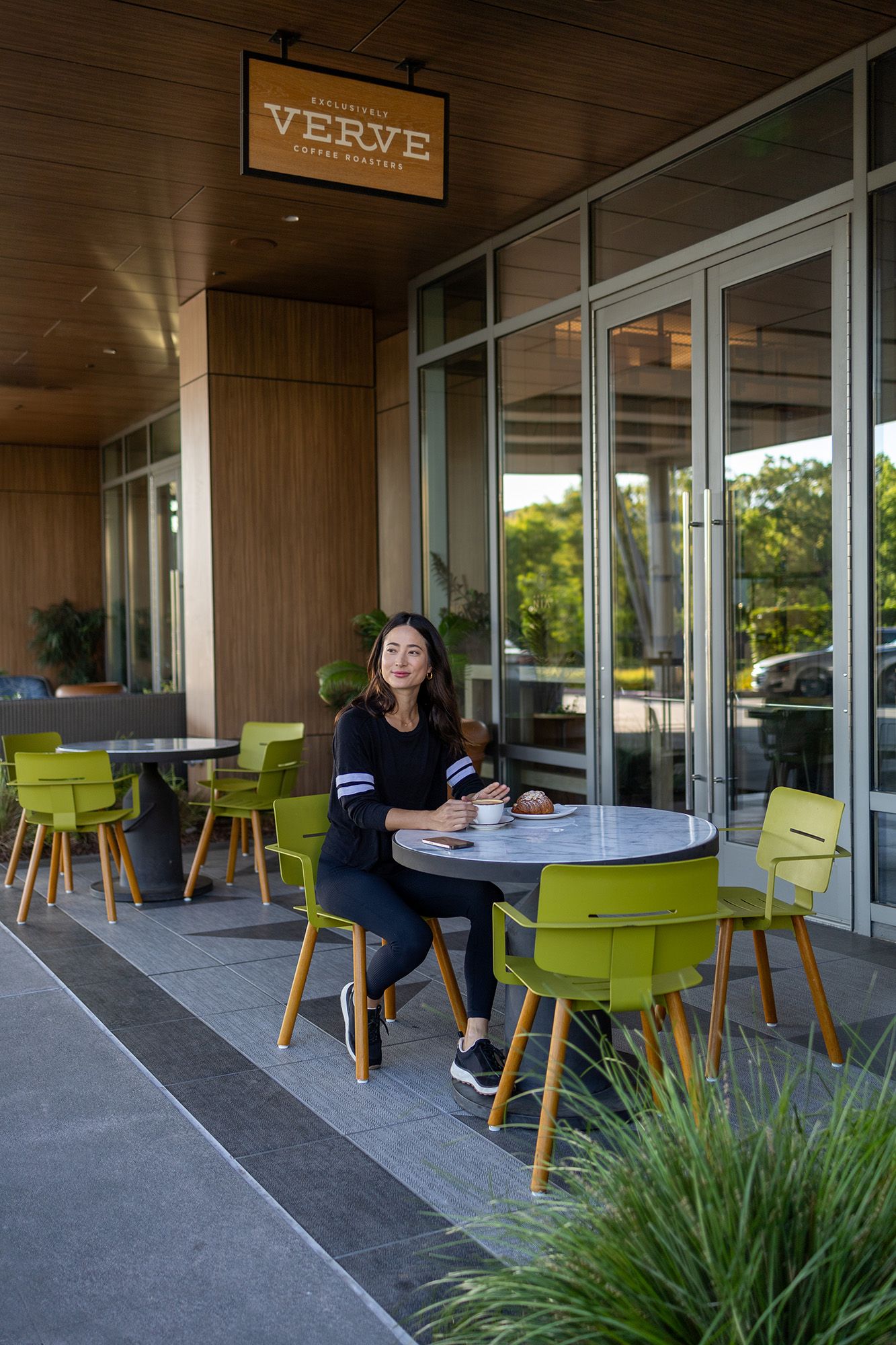 Woman sitting alone at outdoor patio table at Verve Coffee Roasters with green chairs and modern architecture.