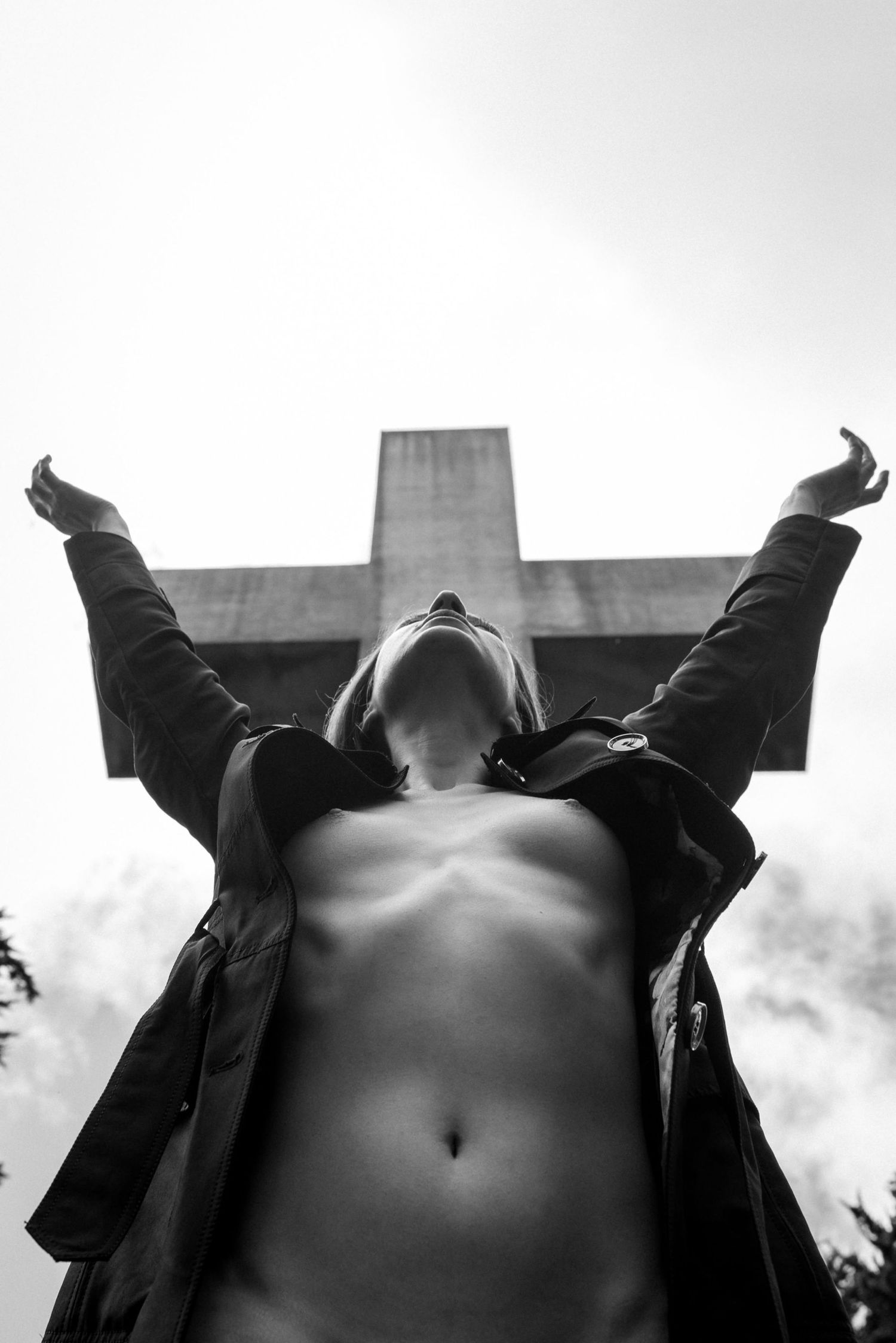 Black and white photo of person with arms raised before a large cross, shot from below against cloudy sky.