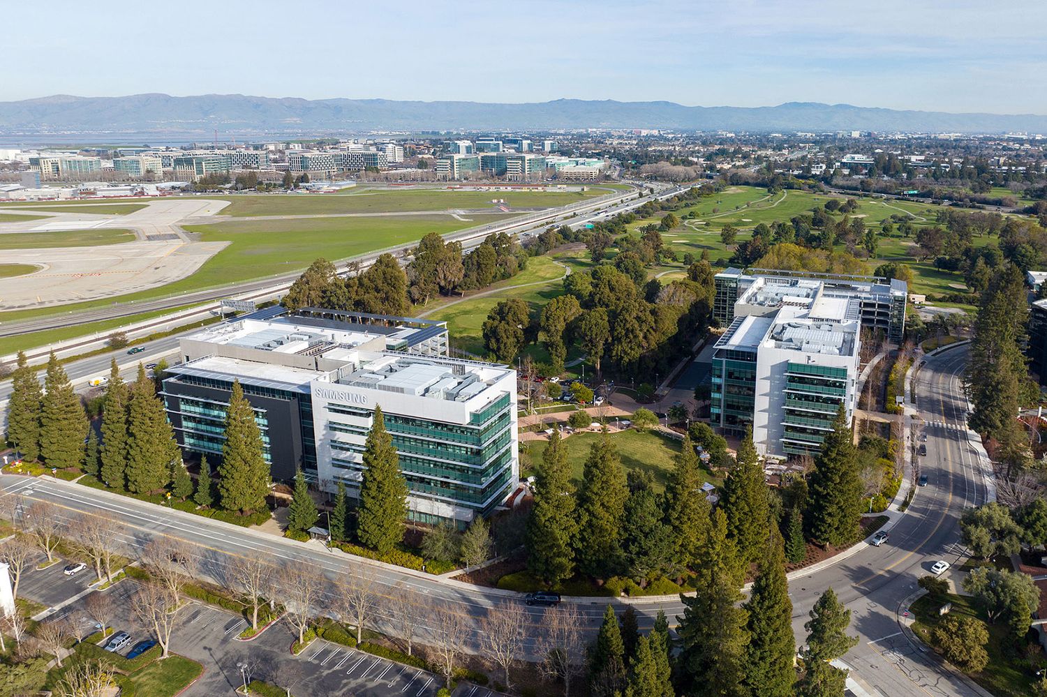Aerial view of modern glass office buildings surrounded by trees near an airport runway.