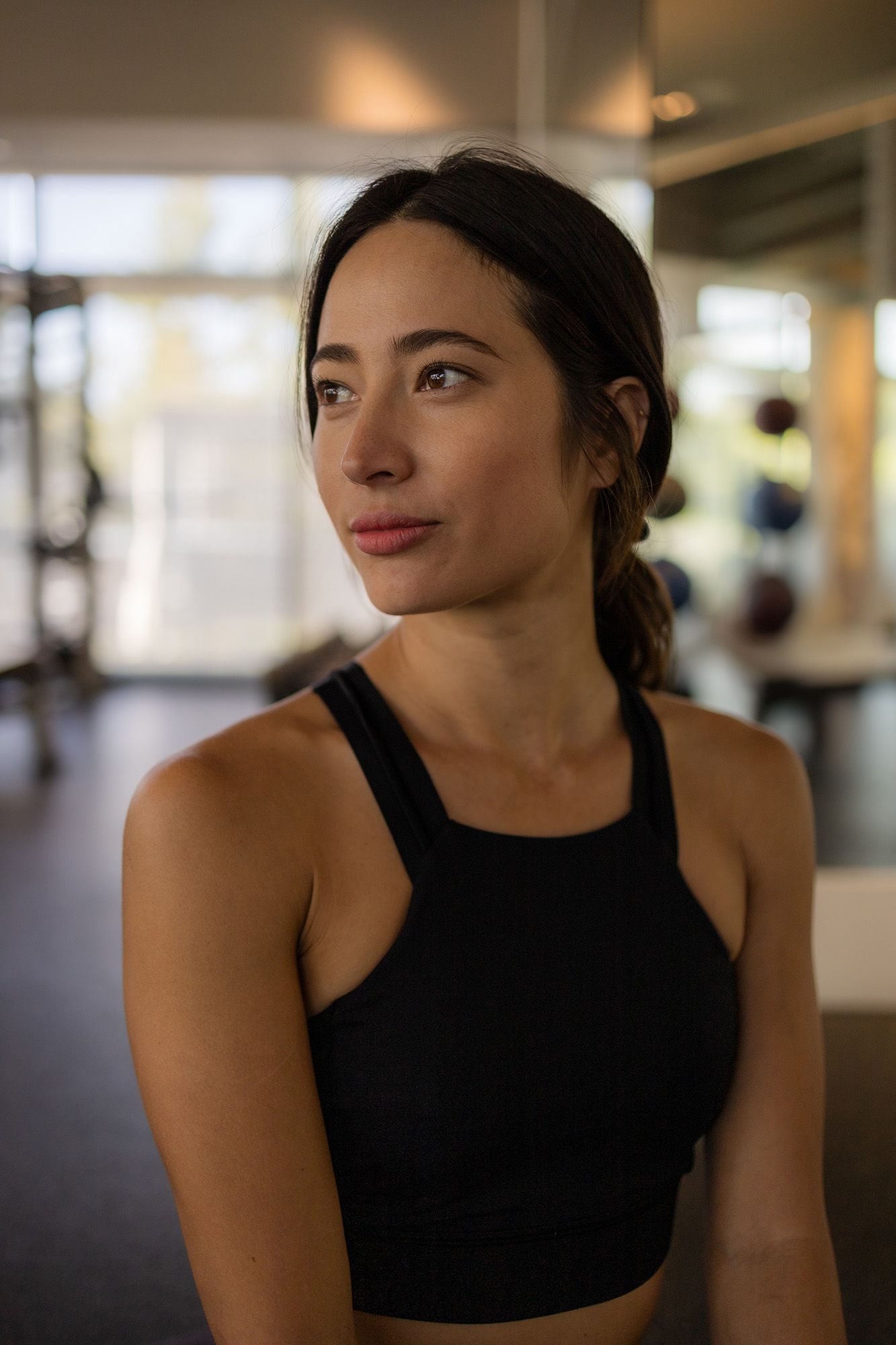 A fit woman in a black sports bra looks away thoughtfully in a modern gym setting.