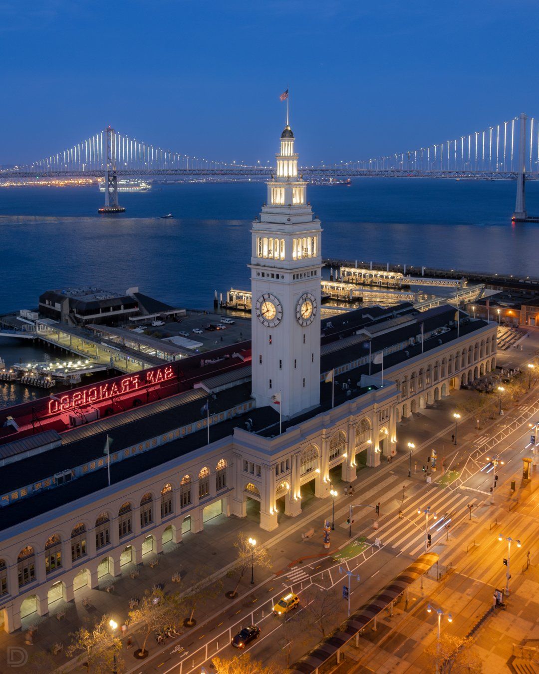 Blue hour aerial view of San Francisco Ferry Building clock tower with Bay Bridge lights reflecting on water.
