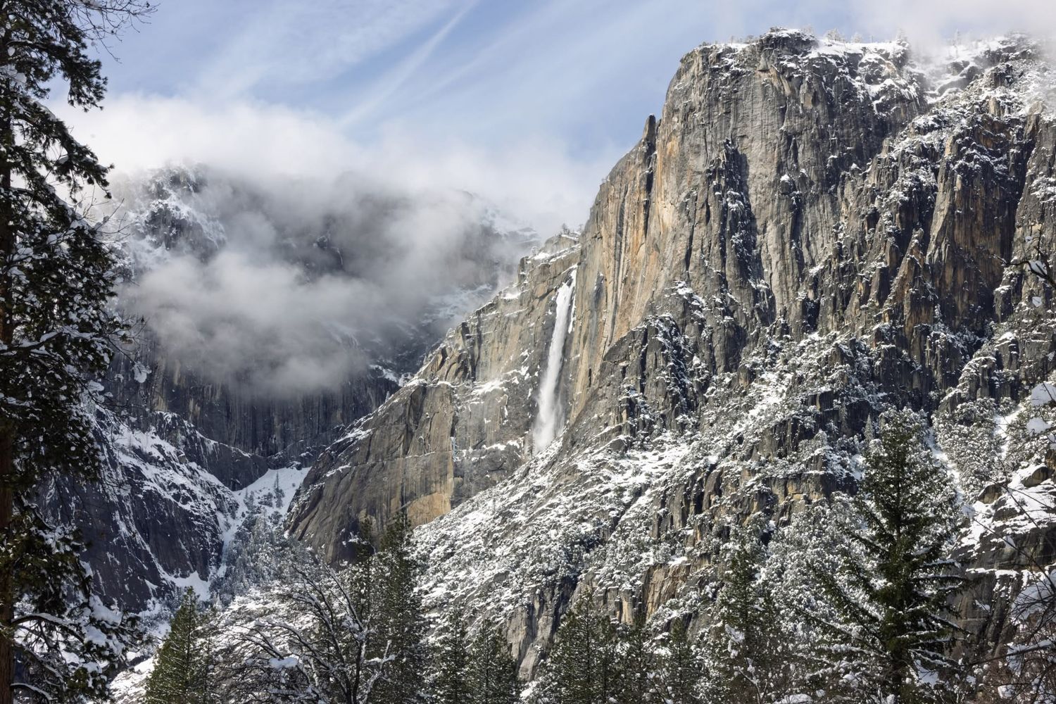 Snow-covered Yosemite Valley cliffs with a waterfall and misty clouds surrounding granite peaks in winter.