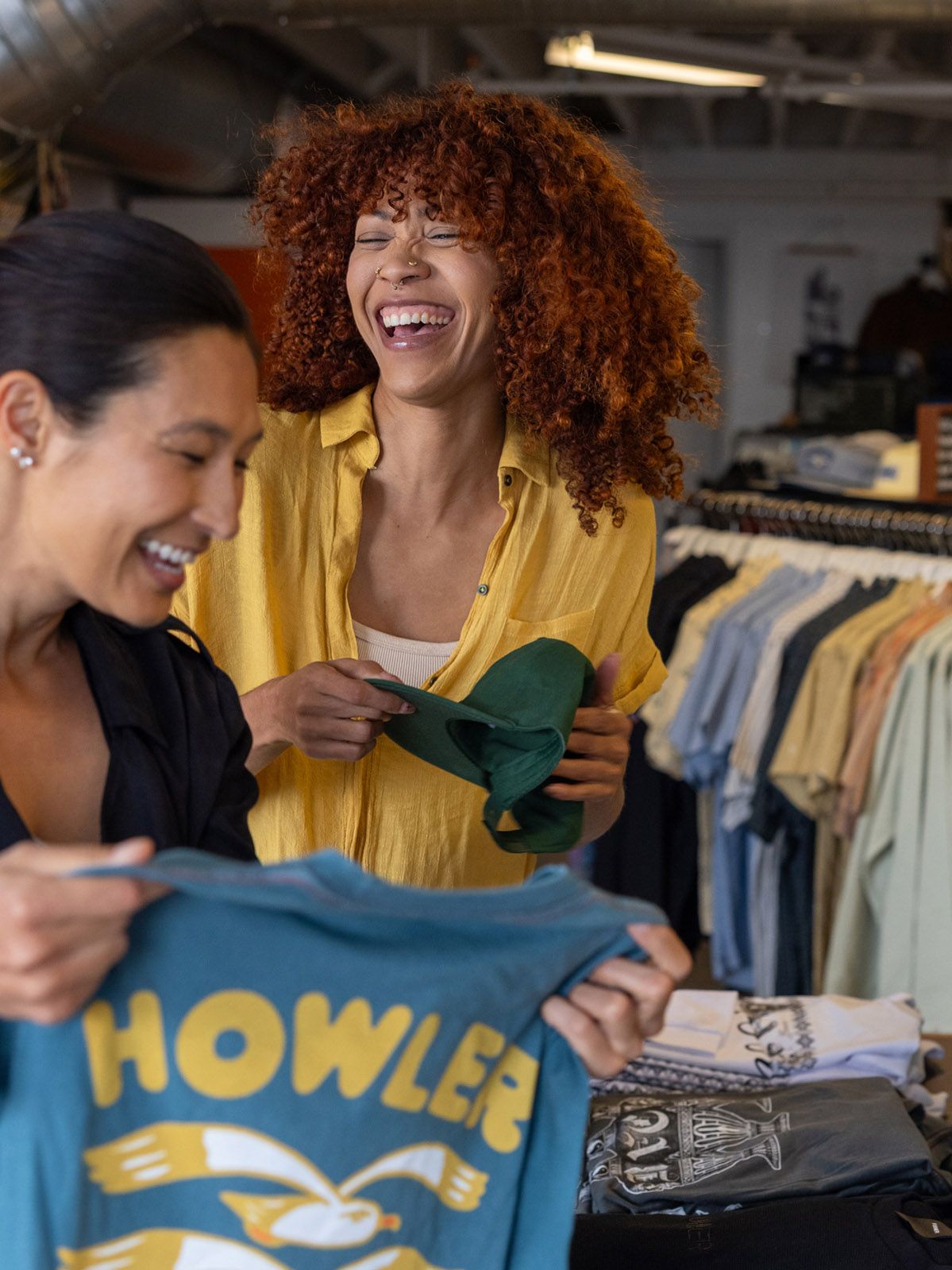 Two women laughing joyfully while browsing colorful graphic tees in a vibrant retail clothing store.