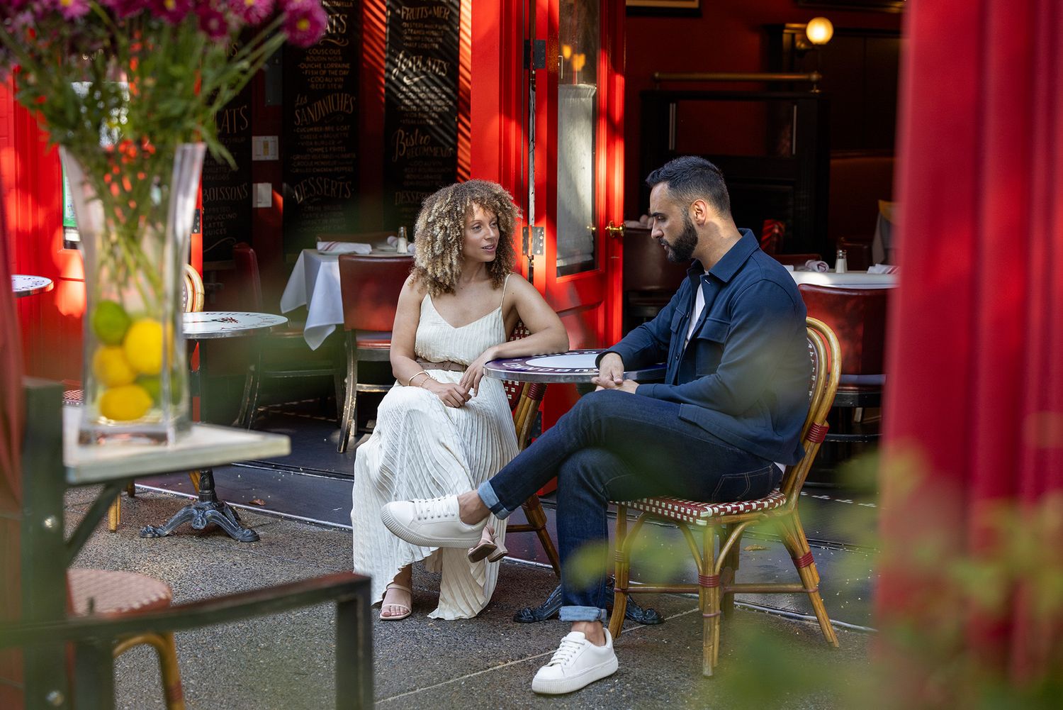 Couple sharing an intimate conversation at a stylish red outdoor cafe patio, woman in white dress, man in navy suit.
