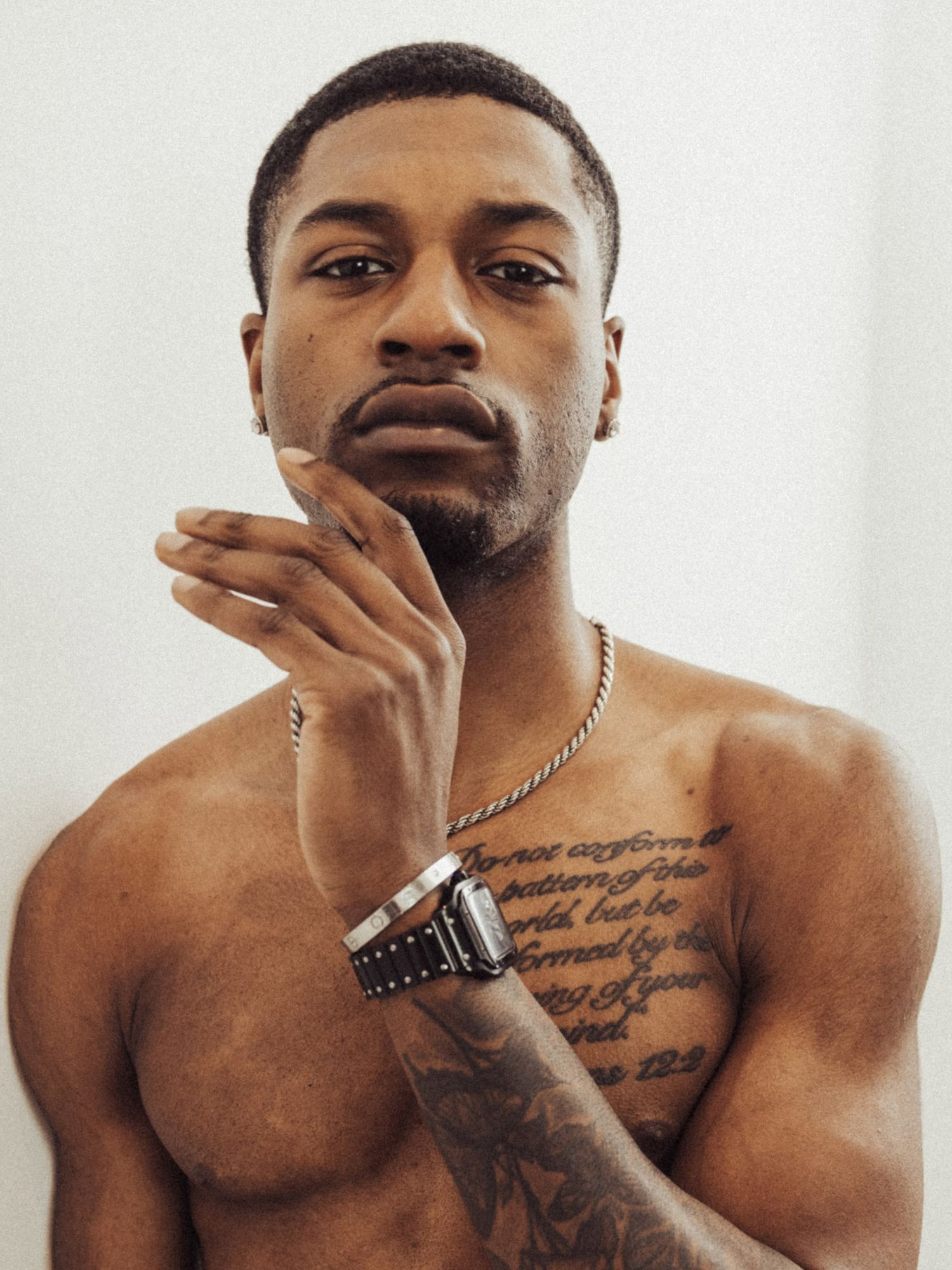 Shirtless man with chest tattoos and chain necklace posing confidently against a white background.