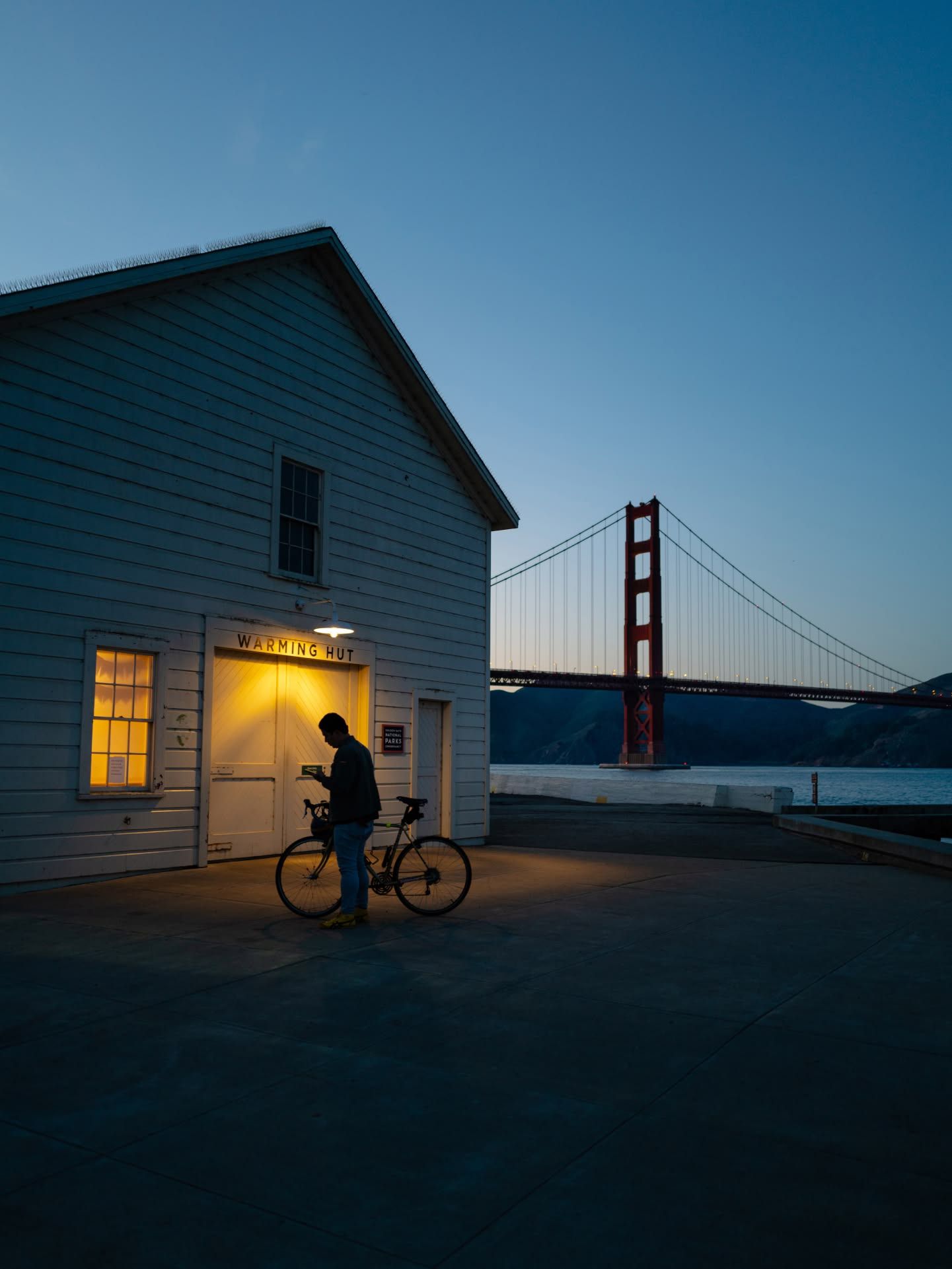 Silhouette of a cyclist near a white building at dusk with the Golden Gate Bridge glowing in the background.
