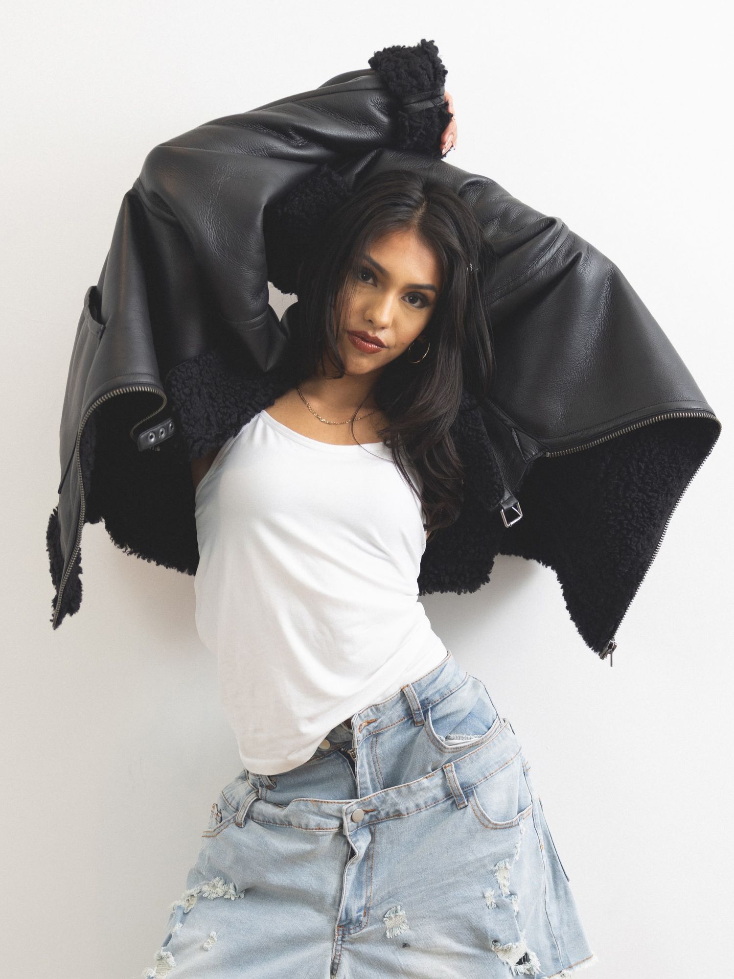 Woman posing in white tee, distressed denim, and black leather shearling jacket raised overhead against white background.