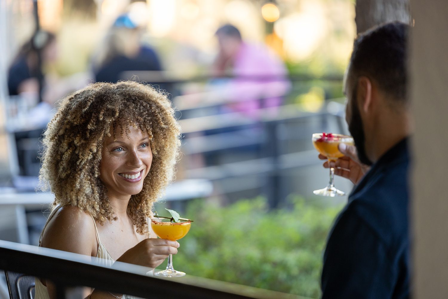 Woman with curly hair smiling and holding a cocktail while enjoying drinks with a friend at an outdoor bar.