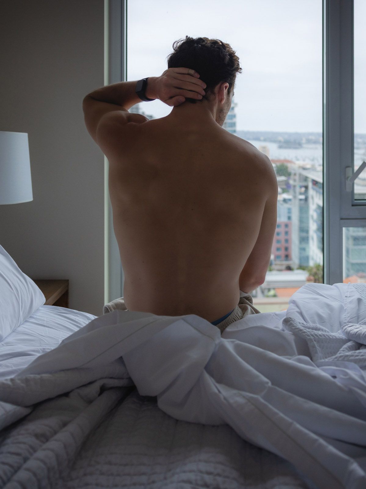 Shirtless man sits on white bed facing city view window in a modern high-rise apartment bedroom.