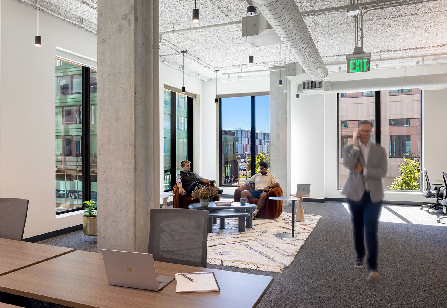Bright contemporary office lounge with concrete columns, pendant lights, and professionals collaborating near large windows.