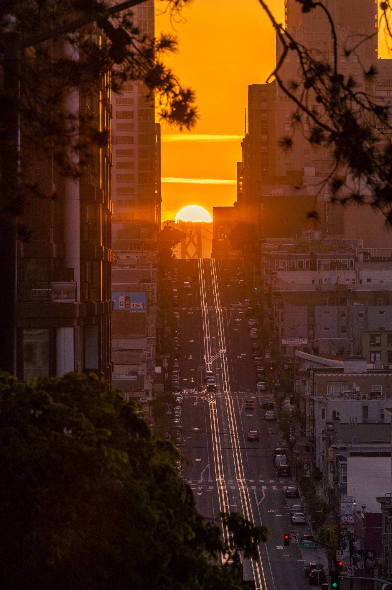 Sun setting perfectly aligned between San Francisco skyscrapers creating a Manhattanhenge effect on a steep street.
