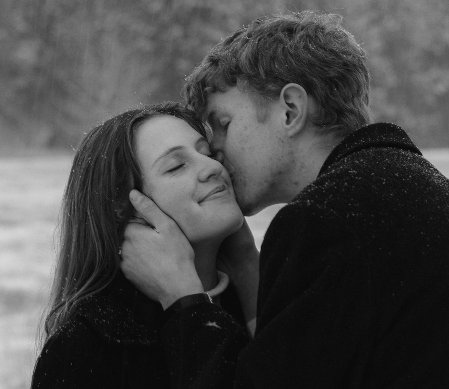 Romantic black-and-white winter portrait of a couple sharing a sweet kiss in a snowy field, capturing timeless love and cozy intimacy.