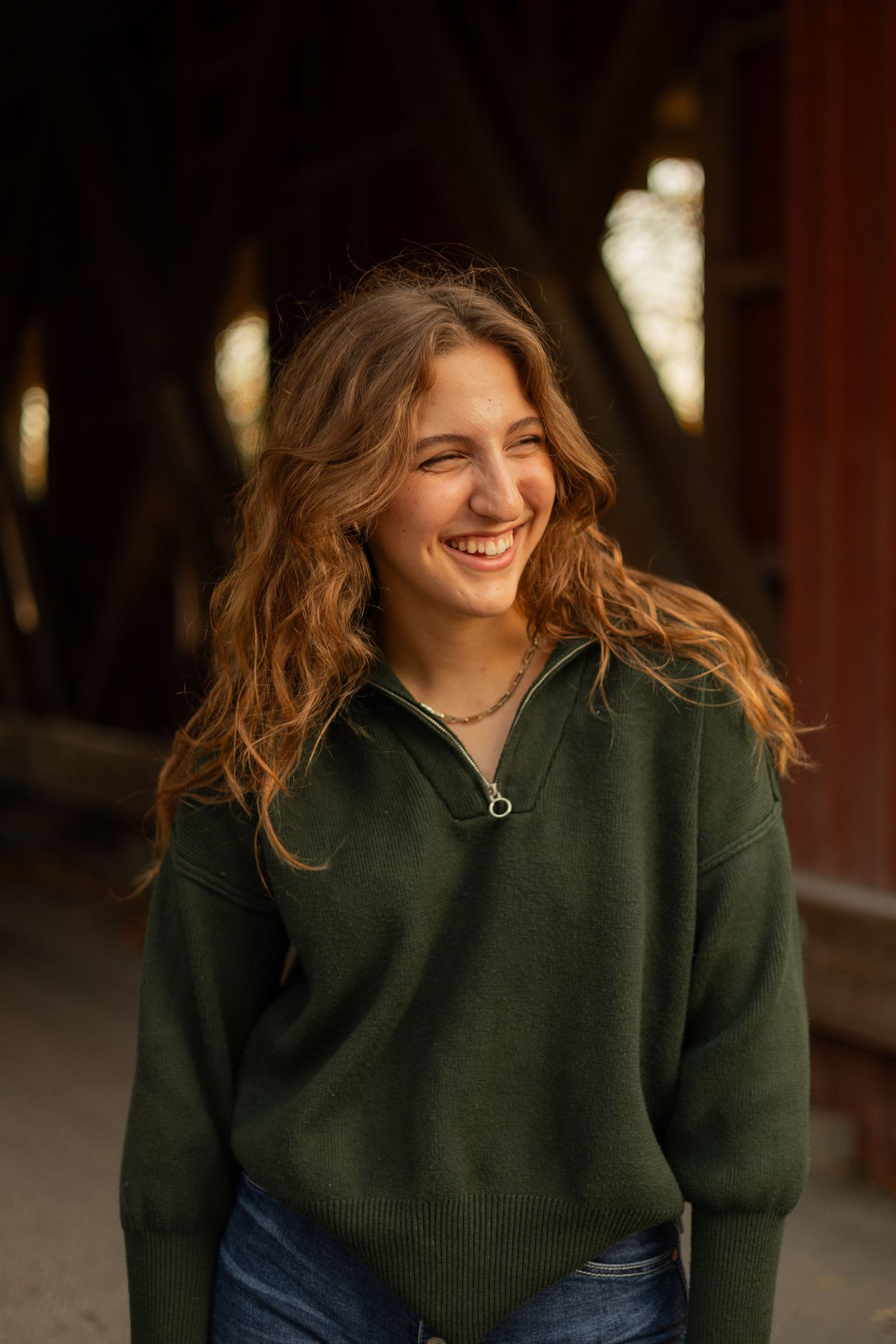 A beautiful woman in a dark green sweater beams with a radiant smile under a rustic bridge, exuding warmth and elegance in golden light.