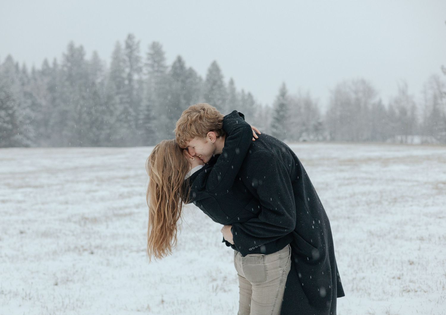A couple embraces in a snowy field, her long hair flowing, him in a sleek black coat—timeless romance captured in a winter wonderland.